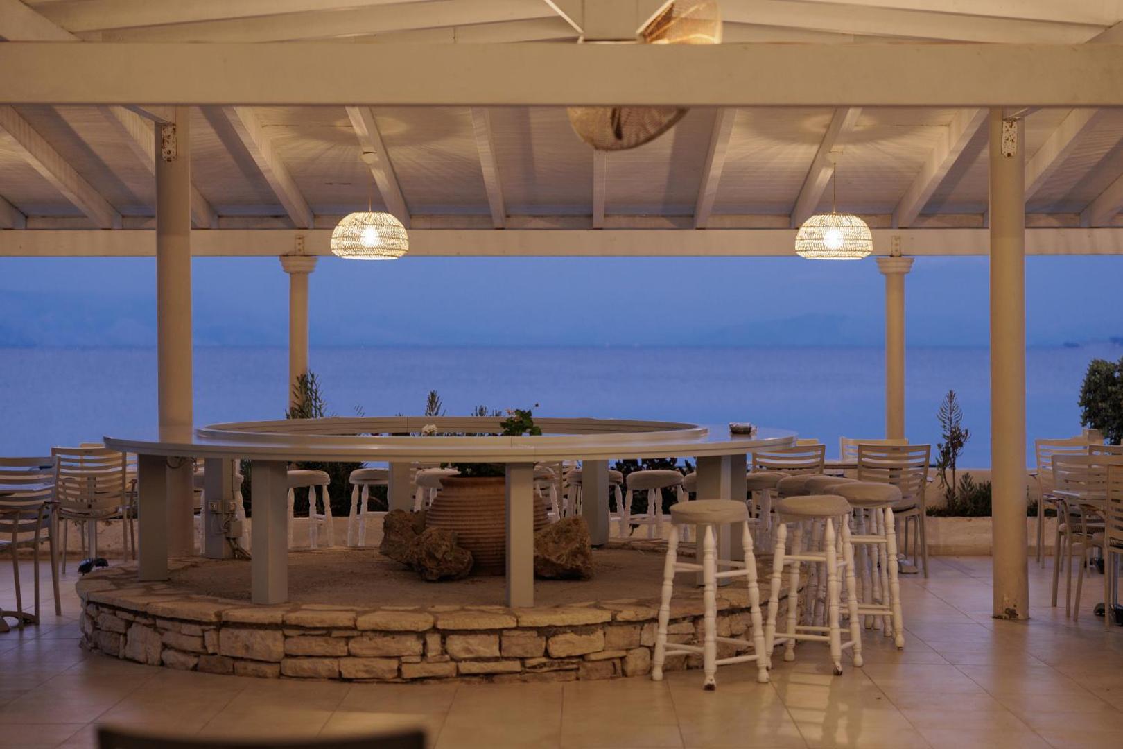 Elea Beach Hotel āra bārs ar skatu uz jūru krēslas stundā, baltiem bāra krēsliem un stilīgu apgaismojumu zem nojumes