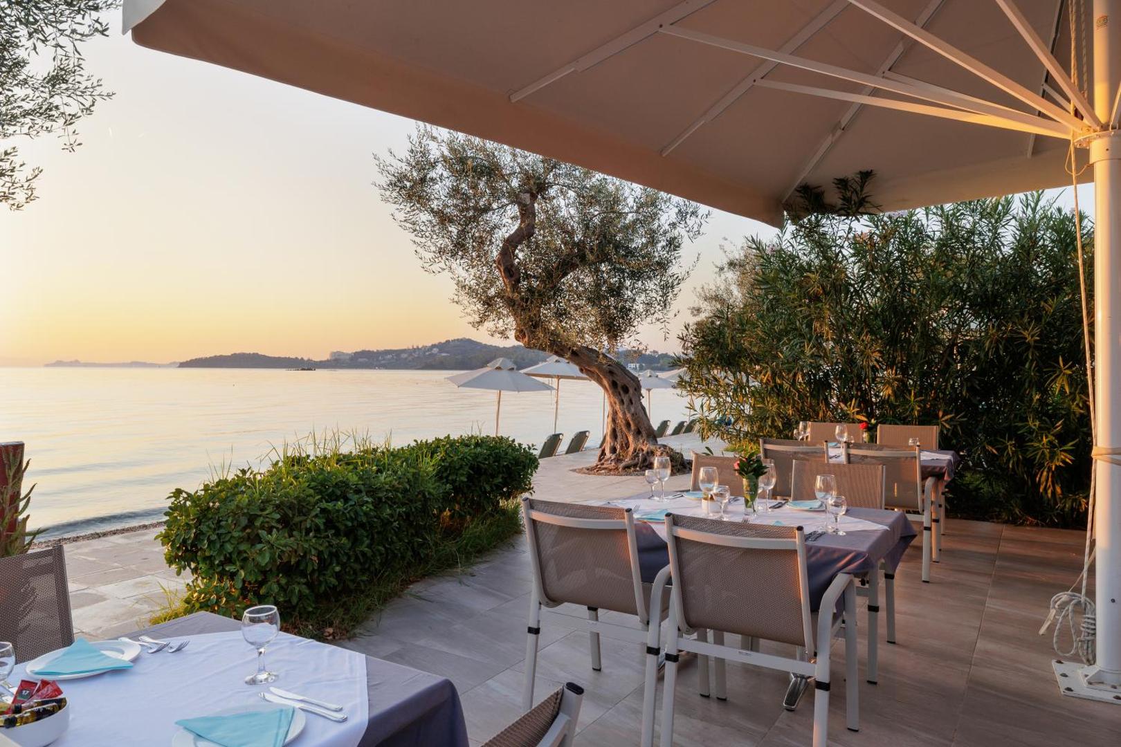 Elea Beach Hotel āra terases restorāns ar galdiem pie jūras saulrietā, apkārtnē augoši olīvkoki un skats uz līci