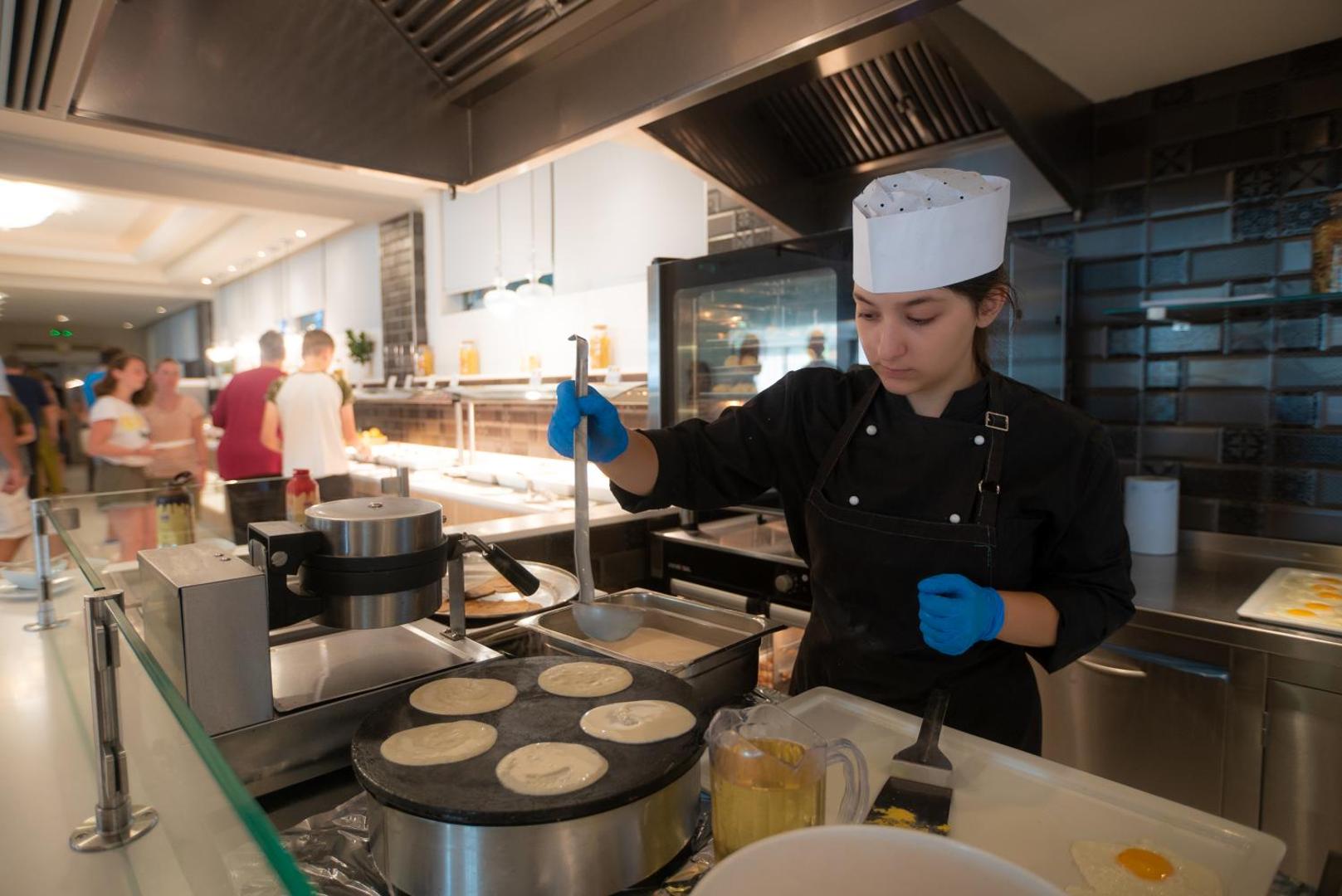 Elea Beach Hotel pavāre gatavo svaigas pankūkas brokastu bufetē, fonā redzami viesi un restorāna interjers