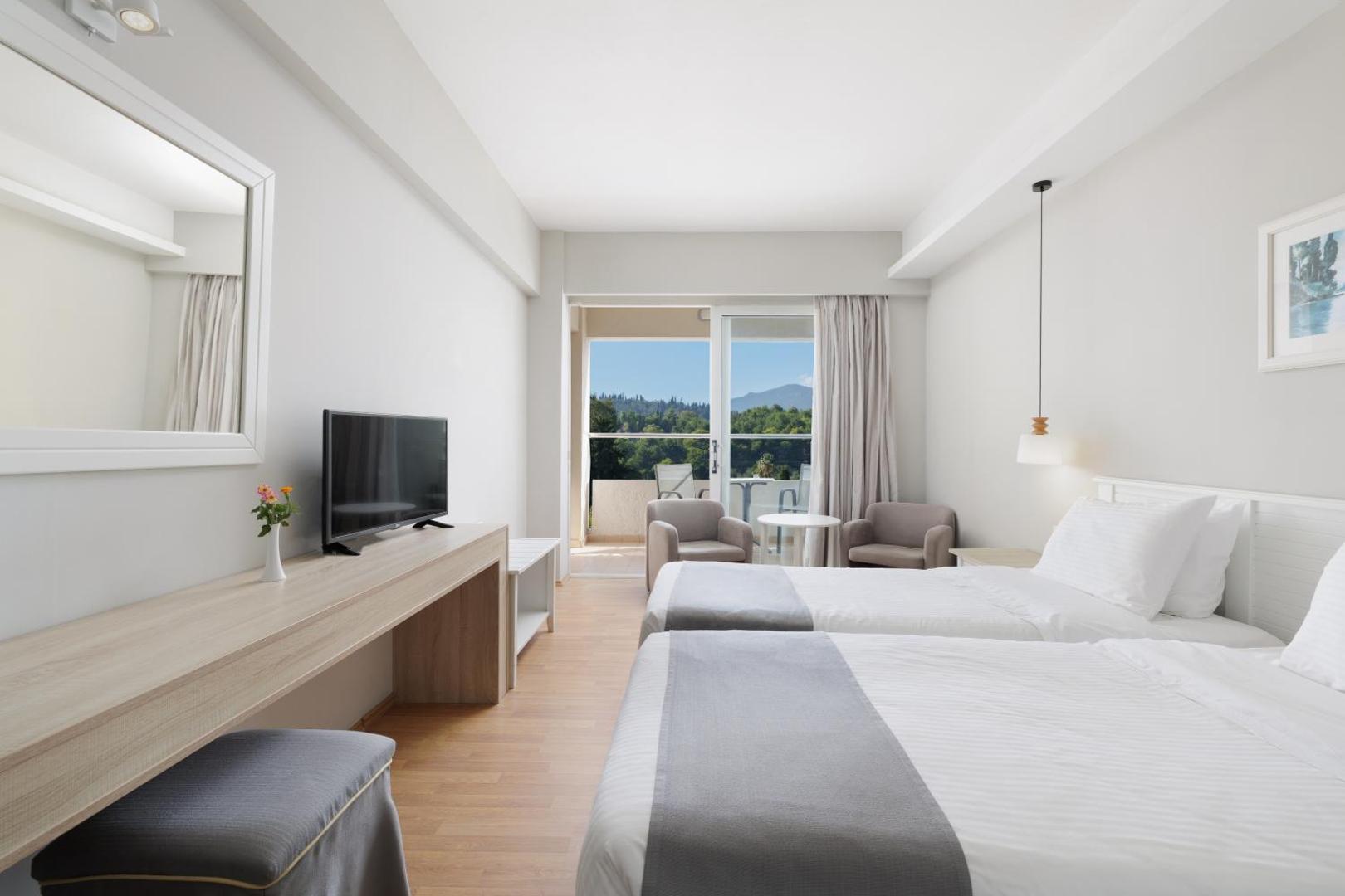 Elea Beach Hotel numura interjers ar skatu uz dārzu un kalniem — rakstāmgalds, televizors un izeja uz balkonu