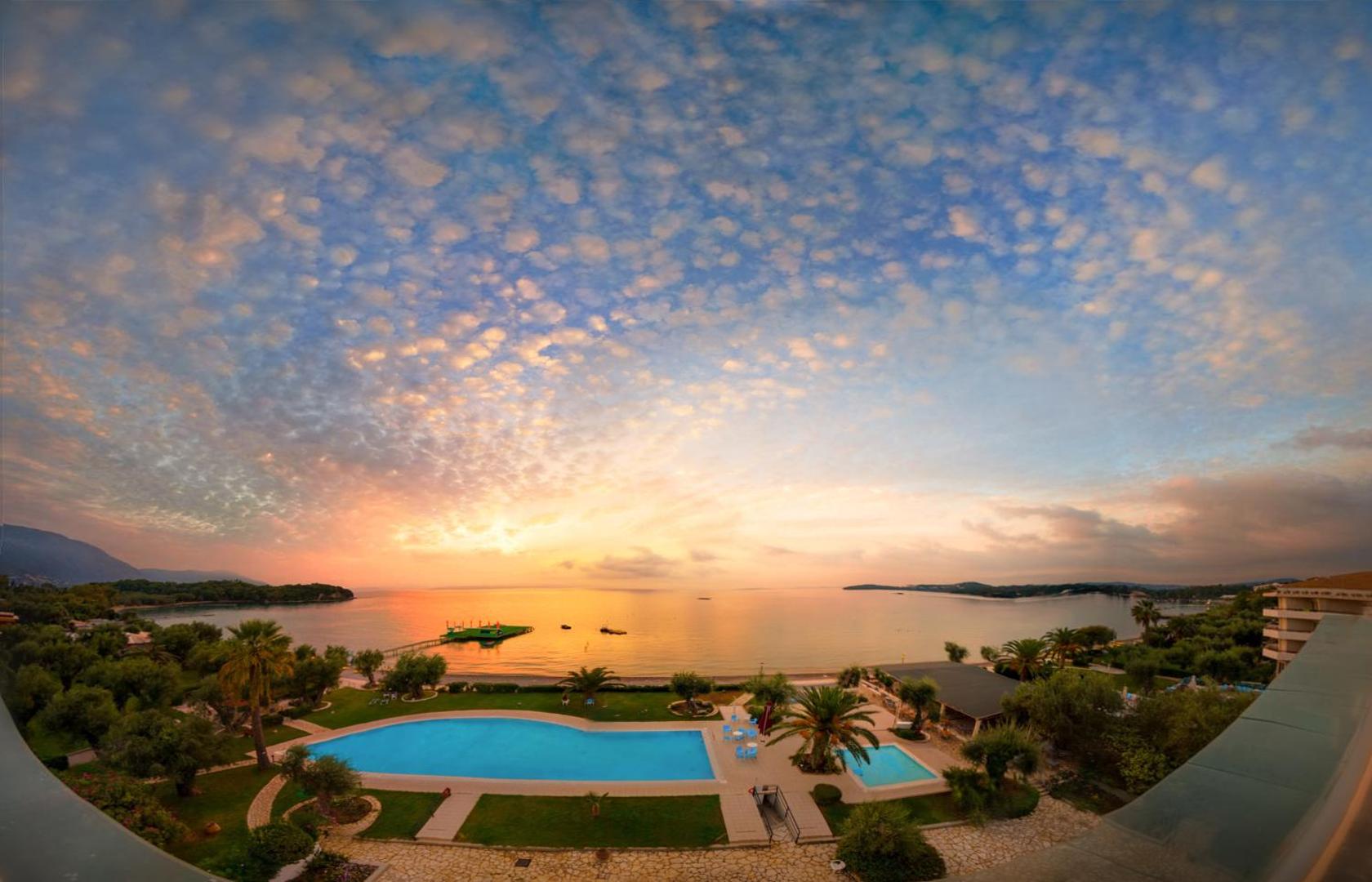 Elea Beach Hotel panorāmas skats uz āra baseinu un Jonijas jūru saulrietā Dasijā, Korfu salā — dārzs ar palmām