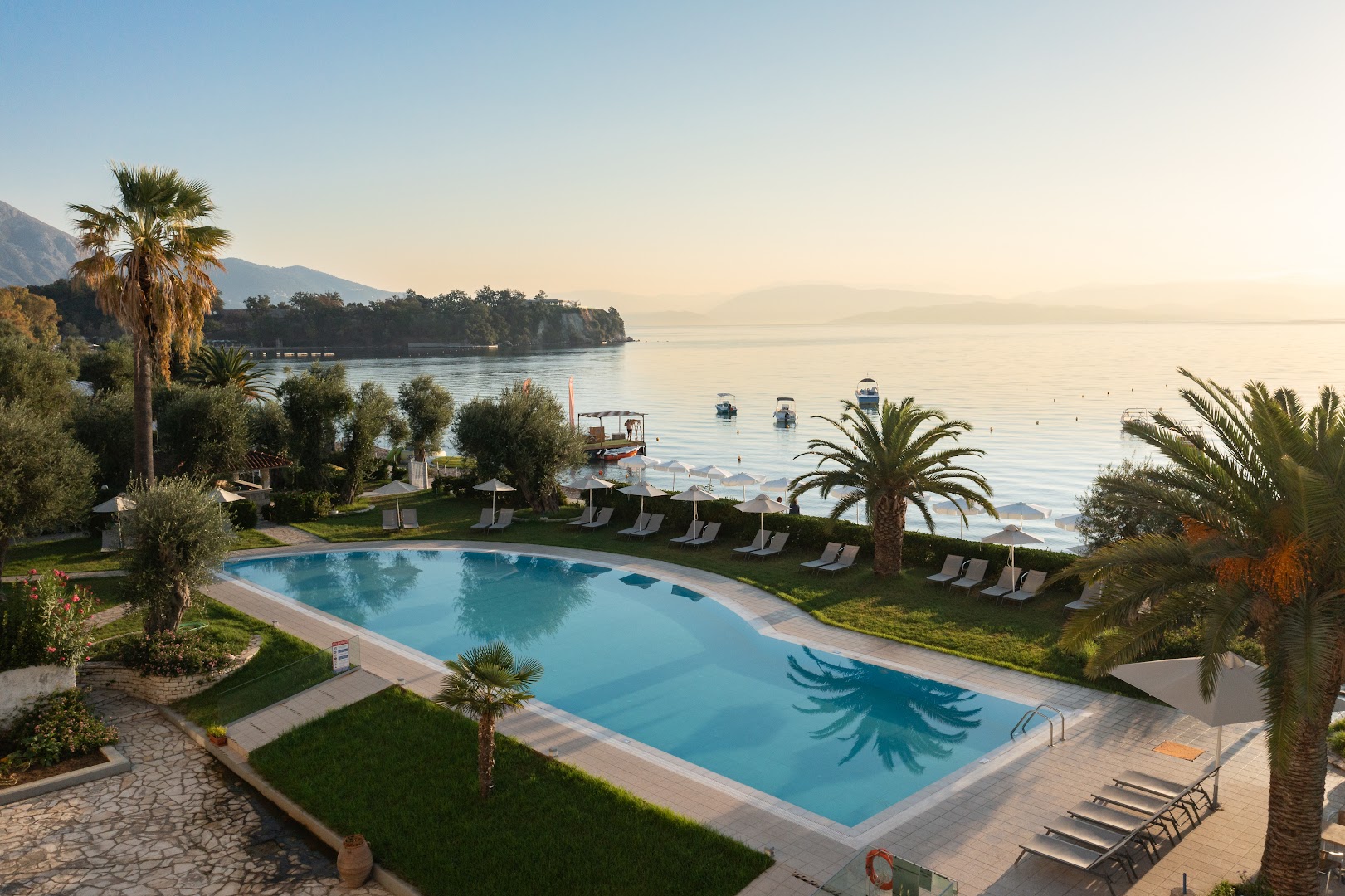 Elea Beach Hotel panorāmas skats uz baseinu, dārzu un Jonijas jūru Dasijā, Korfu — piekrastes ainava ar laivām
