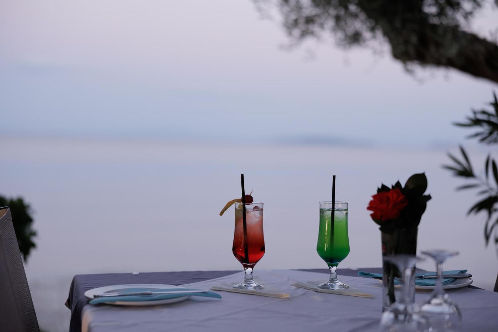 Divi krāsaini kokteiļi uz galda Elea Beach Hotel restorānā piekrastē, fonā redzams mierīgs jūras horizonts saulrietā