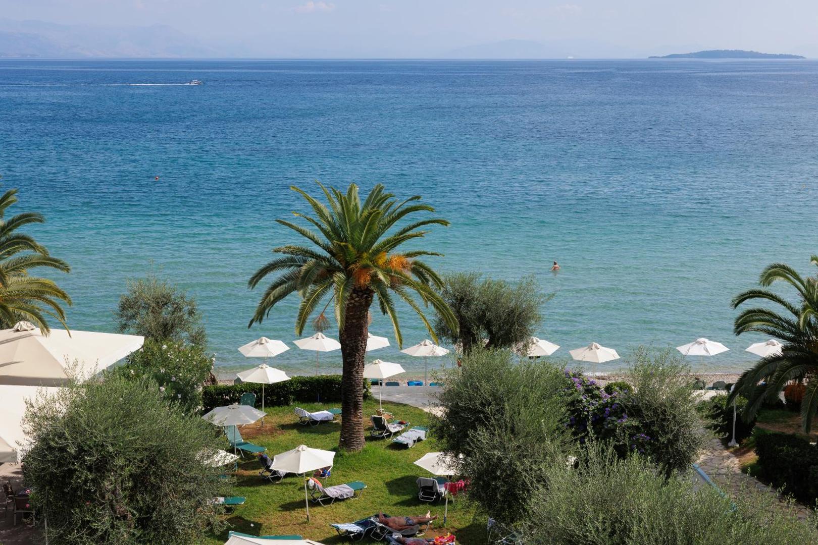 Elea Beach Hotel skats uz pludmali caur palmām un olīvkokiem — zālājs ar saulessargiem un zila jūra fonā