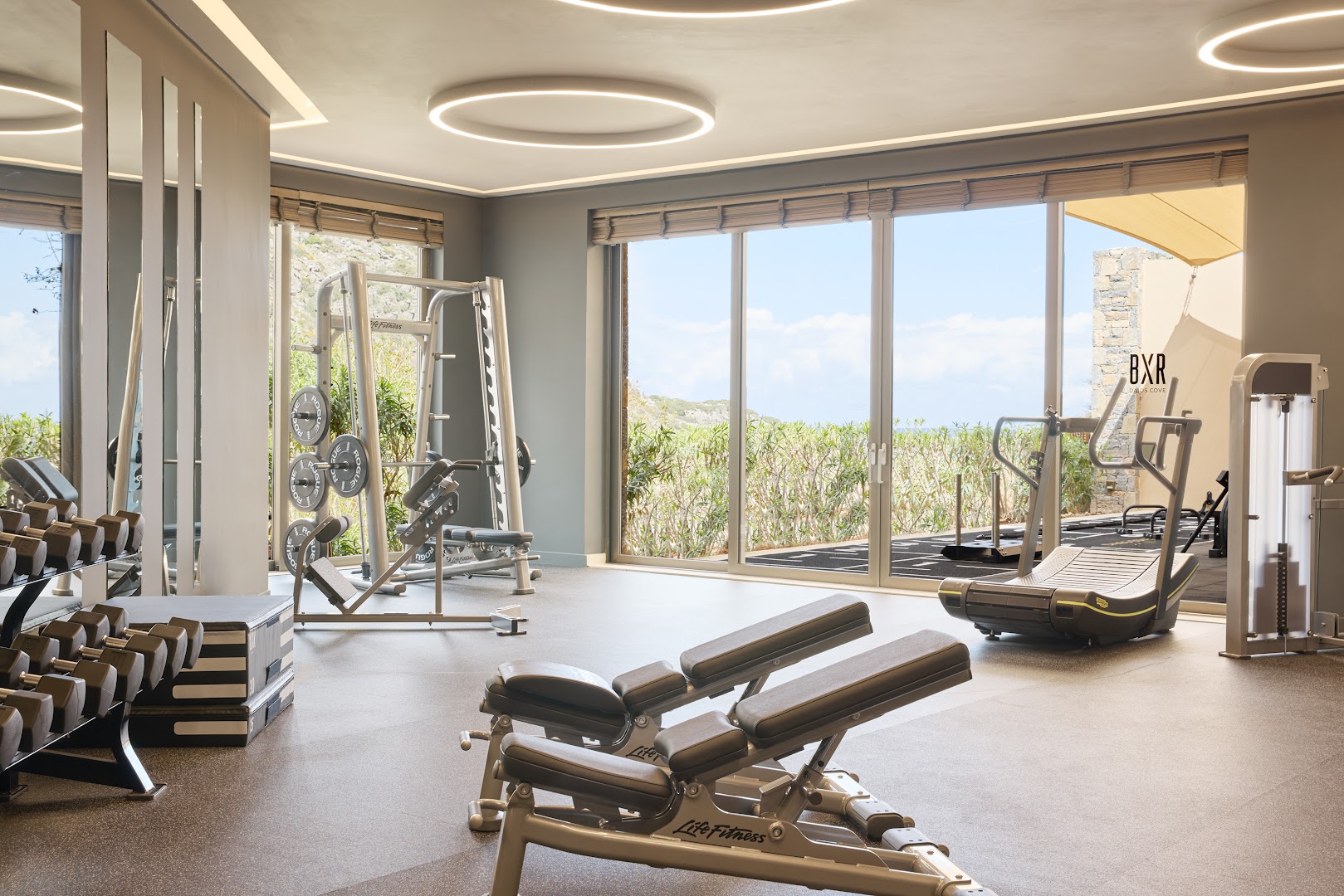 Daios Cove Luxury Resort moderns fitness centrs ar Life Fitness aprīkojumu un panorāmas logiem uz dārzu un jūru