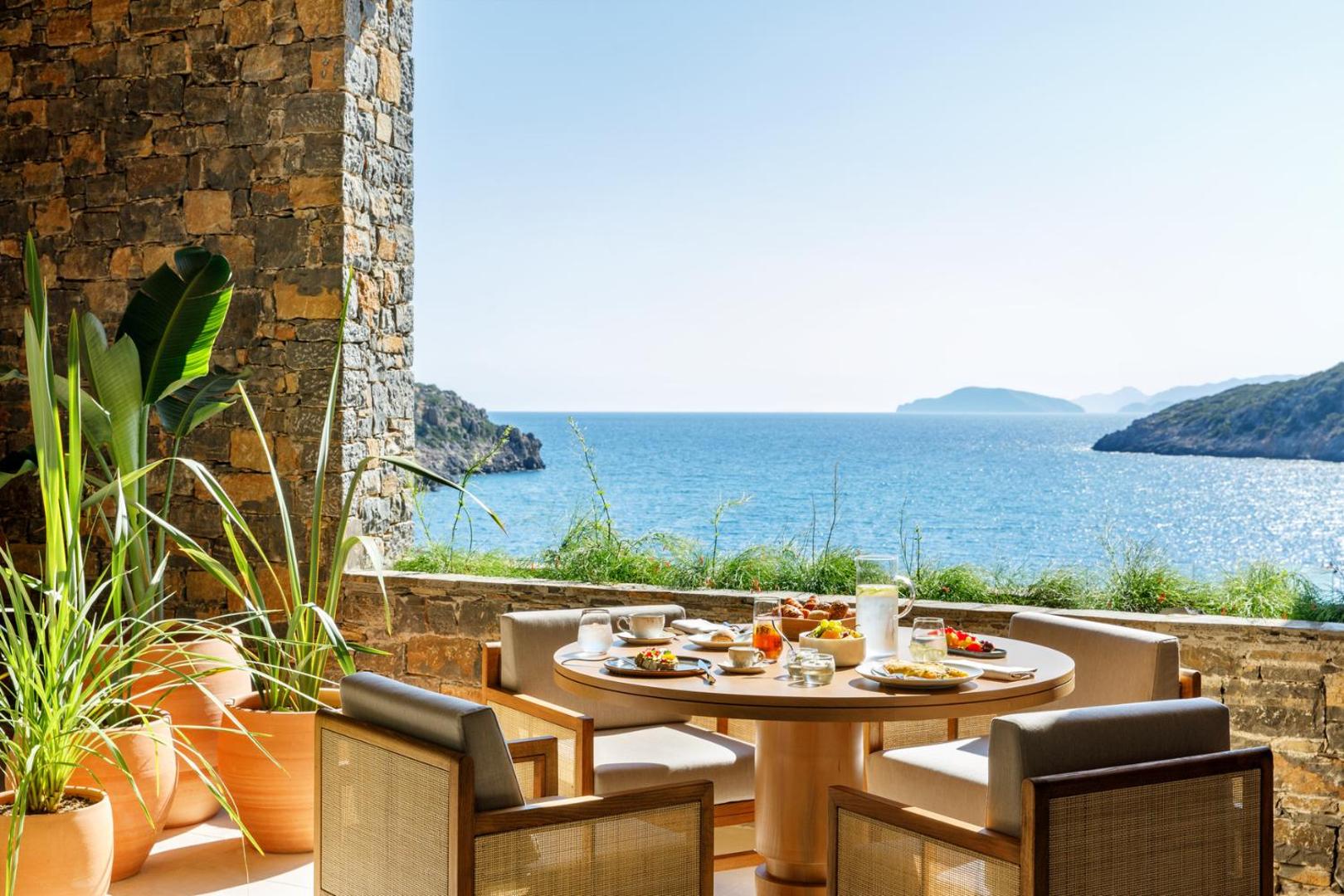 Daios Cove Luxury Resort restorāna terase ar skatu uz Vidusjūru Krētā — klāts brokastu galds pie akmens mūra sienas