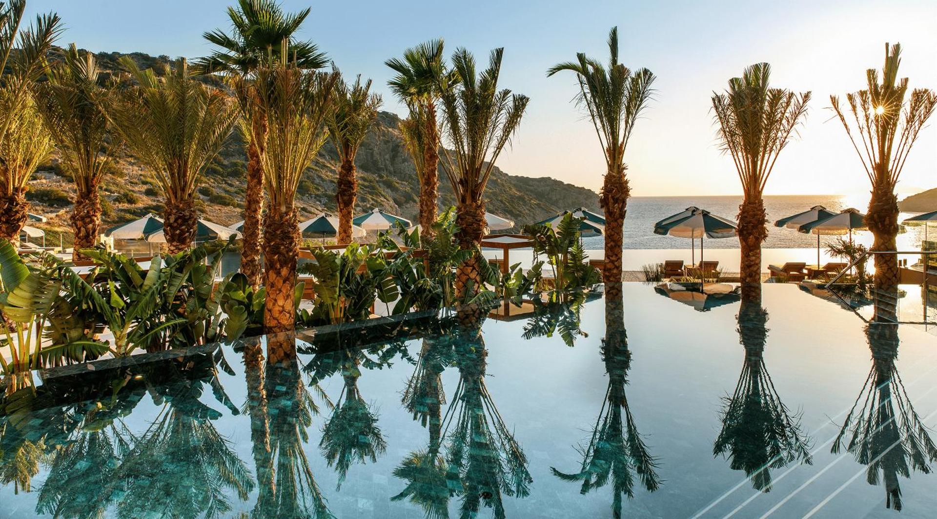 Daios Cove Luxury Resort bezgalības baseins saulrietā ar palmām un skatu uz Vidusjūras līci Krētas piekrastē