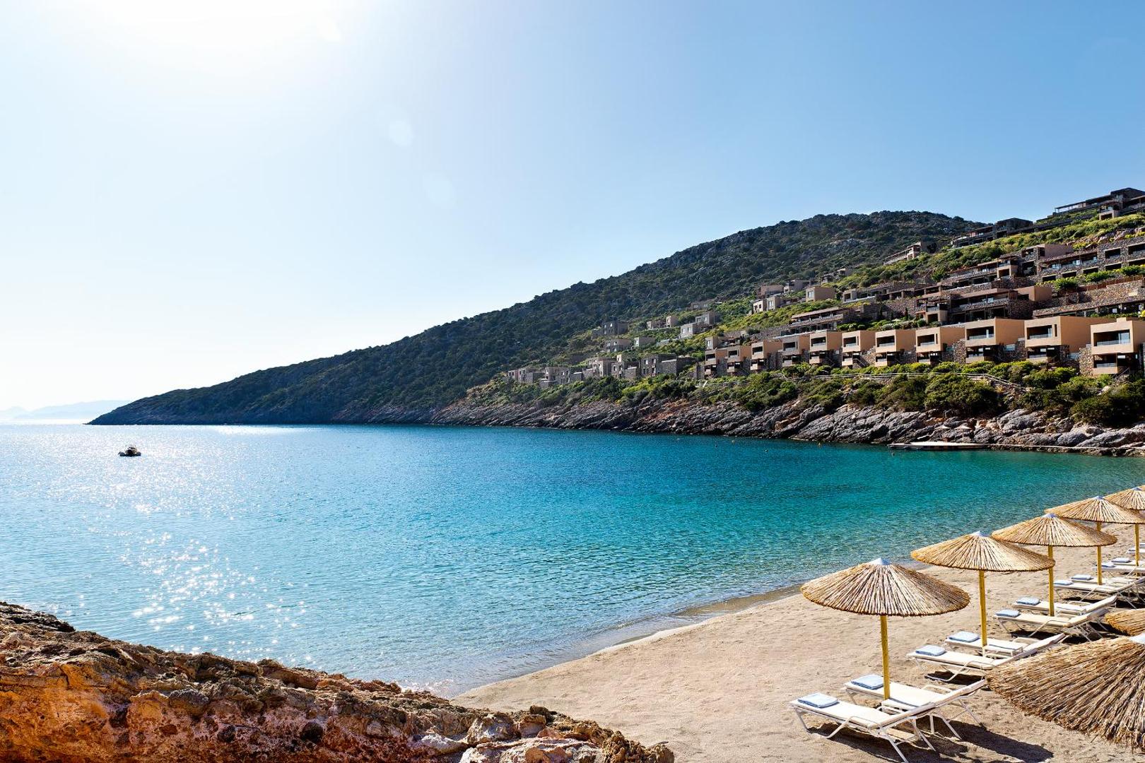 Daios Cove Luxury Resort privātā smilšu pludmale ar saulessargiem un viesnīcas ēkām kalna nogāzē pie tirkīza ūdens