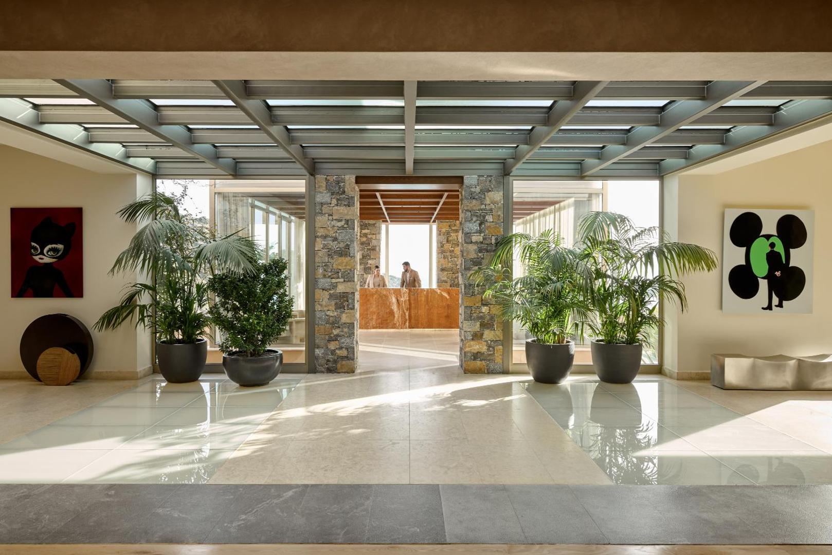 Daios Cove Luxury Resort gaišā recepcijas zona ar stikla jumtu, lieliem telpaugiem un akmens apdari Agios Nikolaos