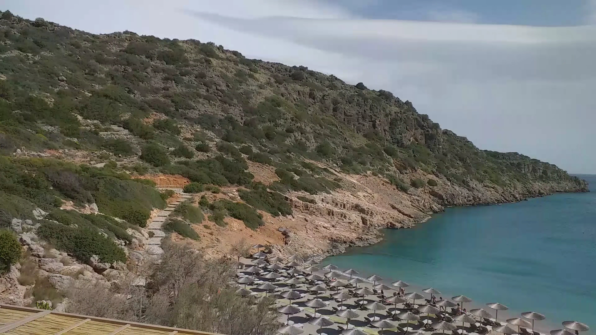 Skats uz Daios Cove Luxury Resort pludmali no augšas ar daudziem saulessargiem un taku, kas ved pa kalna nogāzi