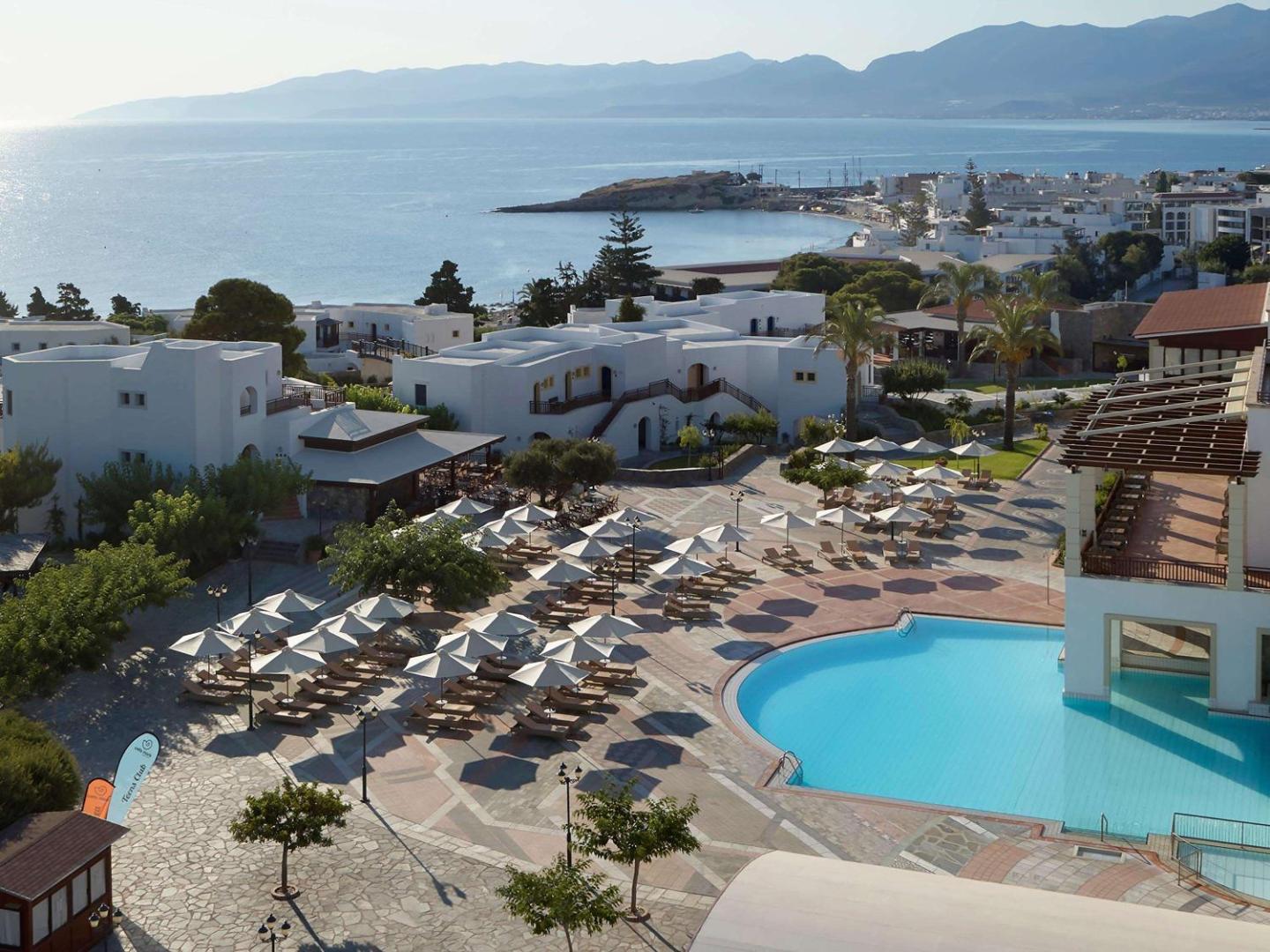 Creta Maris Resort panorāmas skats uz āra baseinu, sauļošanās zonu ar saulessargiem un Hersonisas līci Krētas piekrastē saulainā dienā
