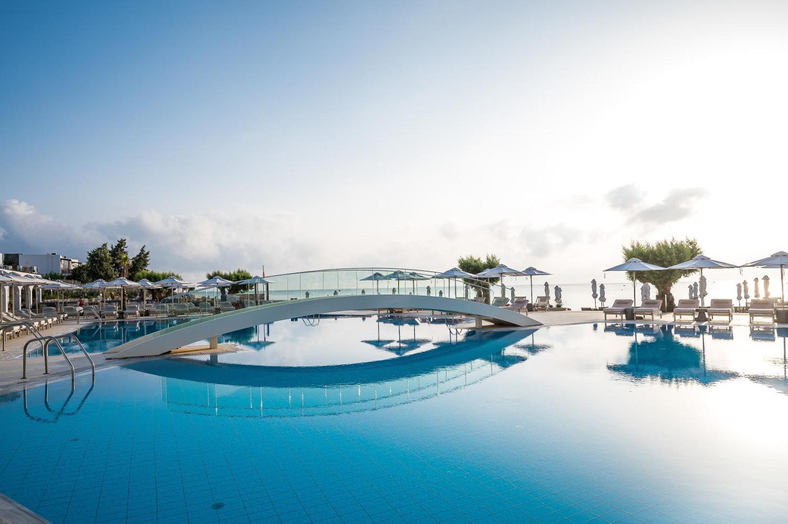 Creta Maris Resort baseins ar dekoratīvu tiltiņu un saulessargiem fonā, skaidras debesis virs Hersonisas piekrastes