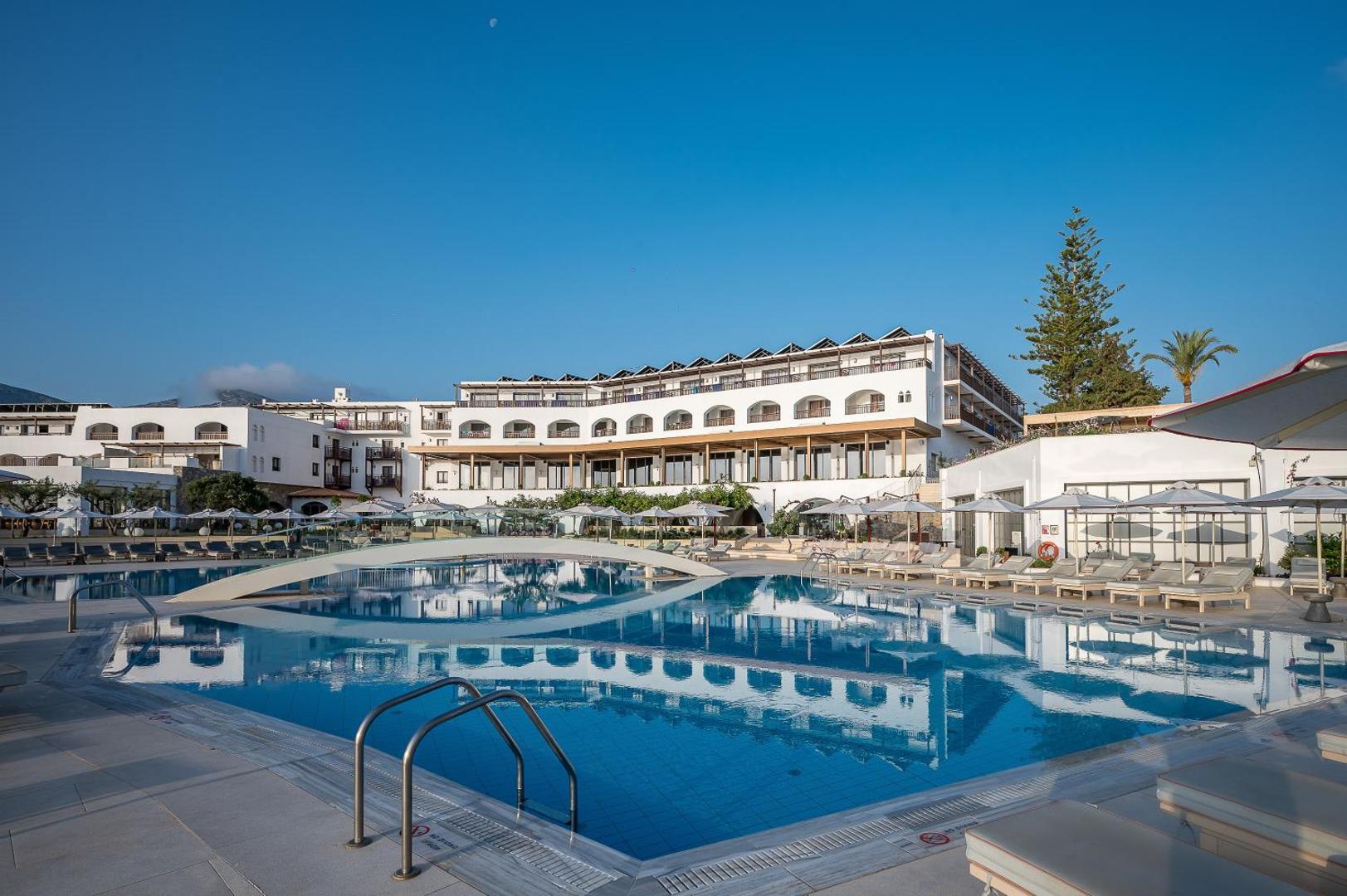 Creta Maris Resort galvenā ēka ar baltu fasādi un lielu āra baseinu, kura ūdenī atspīd viesnīcas arhitektūra