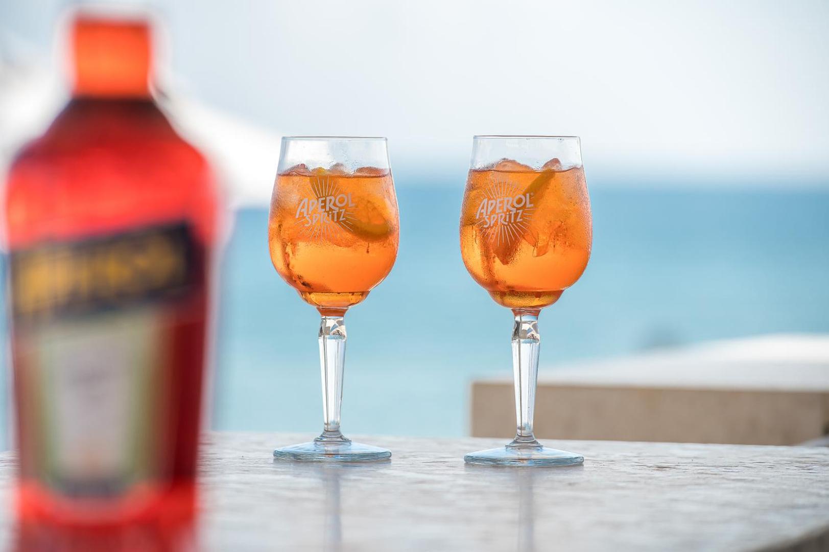 Divi Aperol Spritz kokteiļi uz galda Creta Maris Resort piekrastes bārā ar izpludinātu jūras fonu
