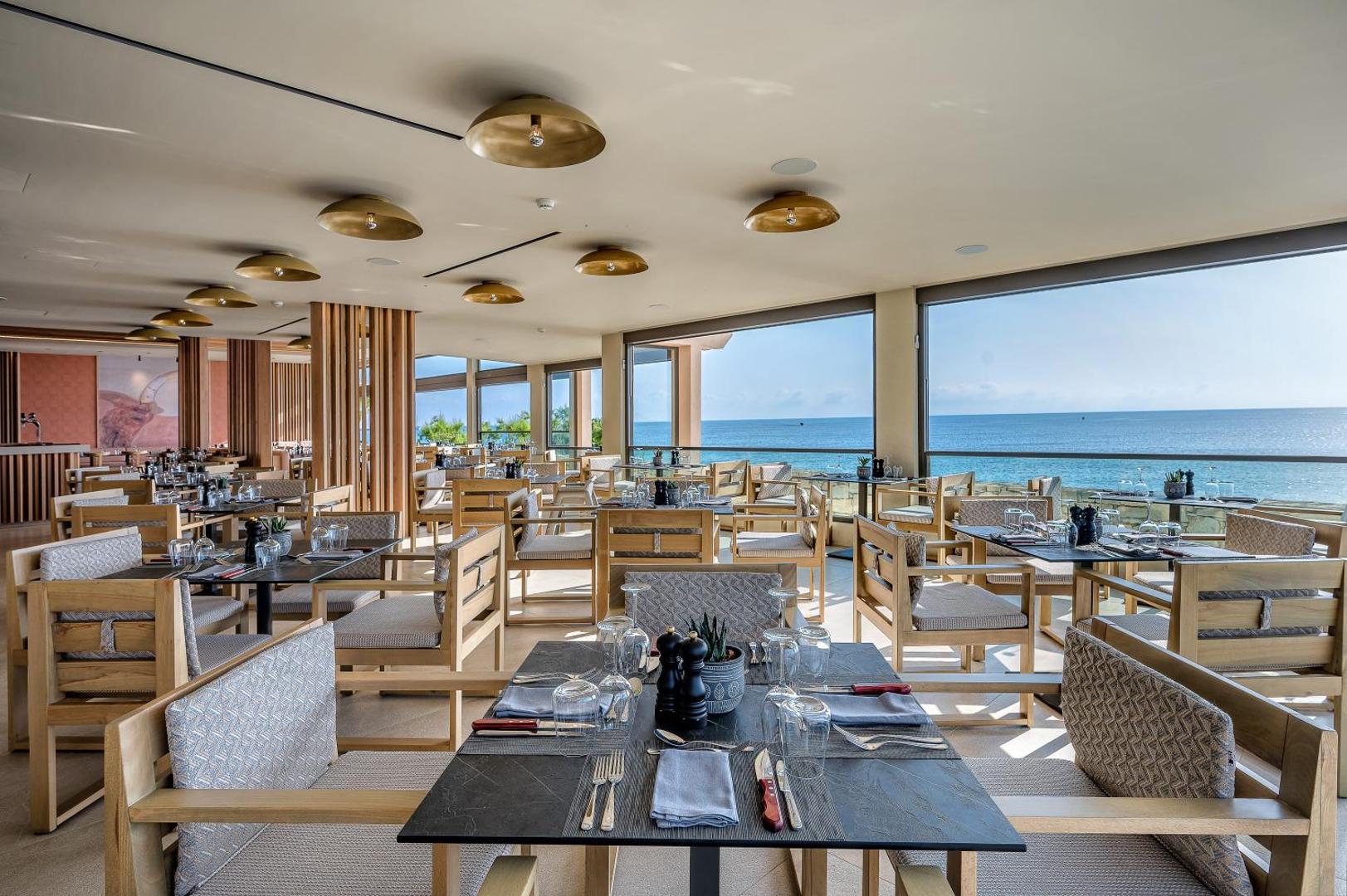 Creta Maris Resort restorāns ar koka mēbelēm un lieliem panorāmas logiem, no kuriem paveras skats uz jūru