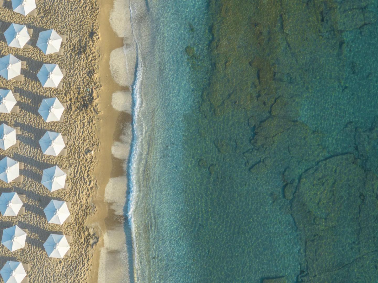 Creta Maris Resort privātā smilšu pludmale no putna lidojuma — balti saulessargi un dzidrs, tirkīzzils ūdens Krētas salā