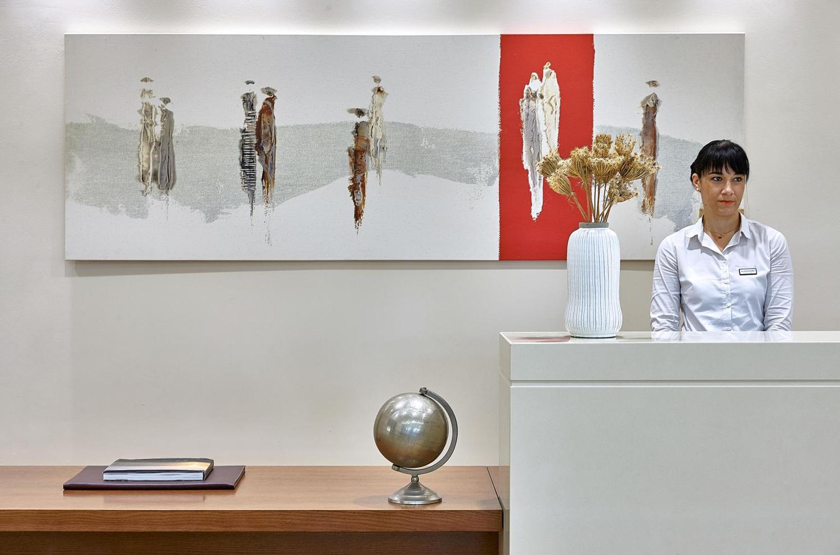 Viesnīcas Castello Boutique Resort & Spa recepcijas zona ar modernu mākslas darbu pie sienas un viesmīlīgu darbinieci pie letes