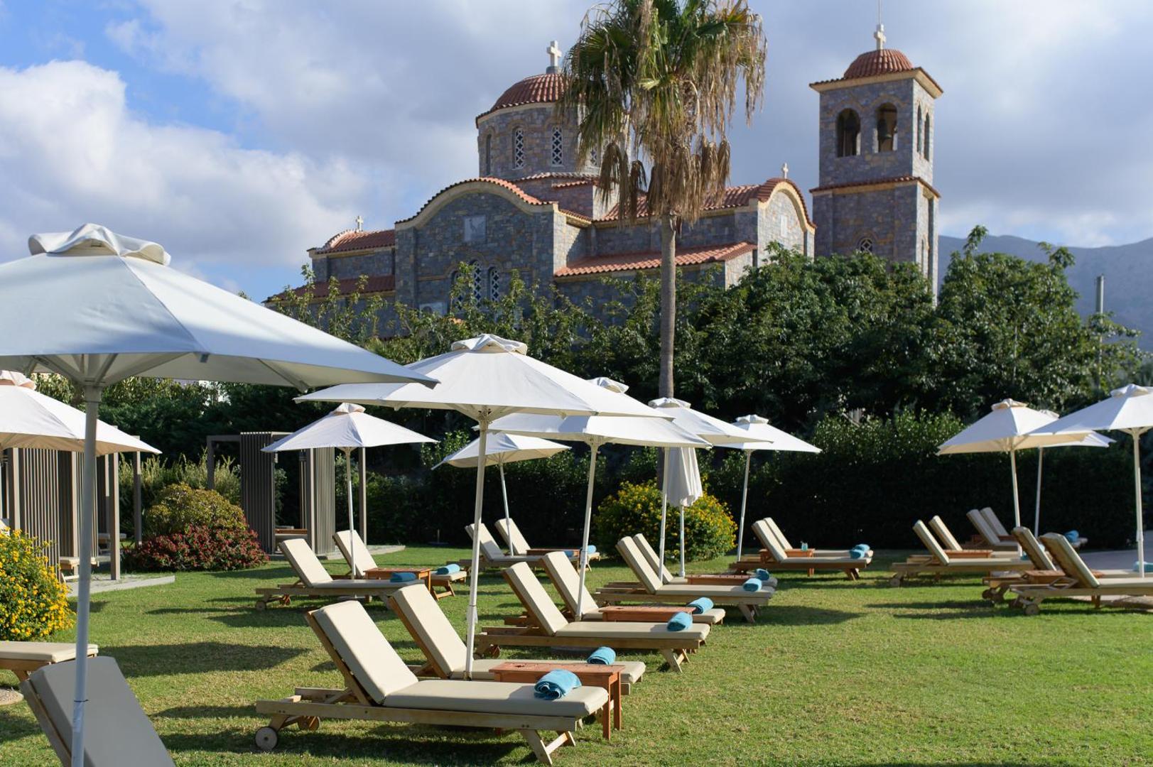 Castello Boutique Resort & Spa atpūtas zona zālienā ar sauļošanās krēsliem un saulessargiem uz vēsturiskas baznīcas fona Krētā