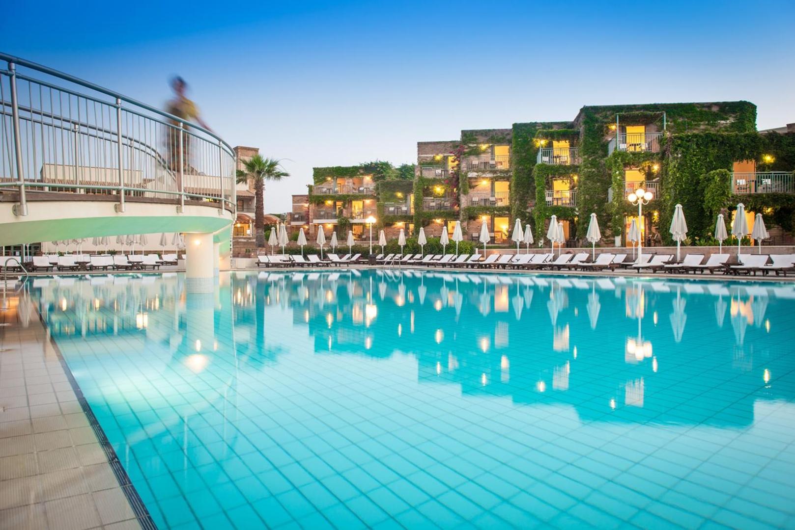 Bella Beach Hotel izgaismots āra baseins vakarā ar skatu uz apstādījumiem klāto viesnīcas fasādi un tiltiņu