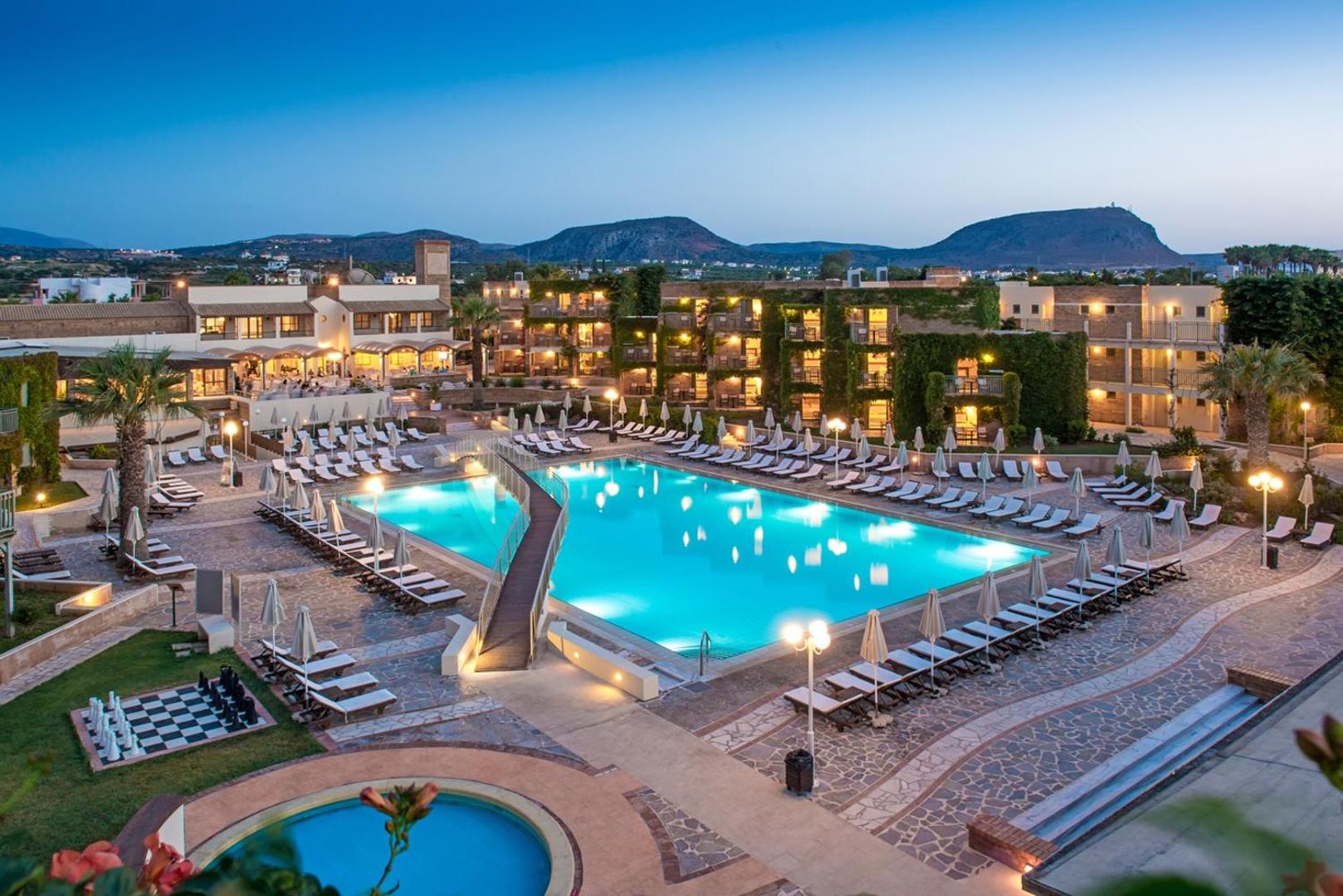 Bella Beach Hotel vakara skats uz izgaismotu āra baseinu, sauļošanās zonu un viesnīcas ēkām uz kalnu fona Krētā