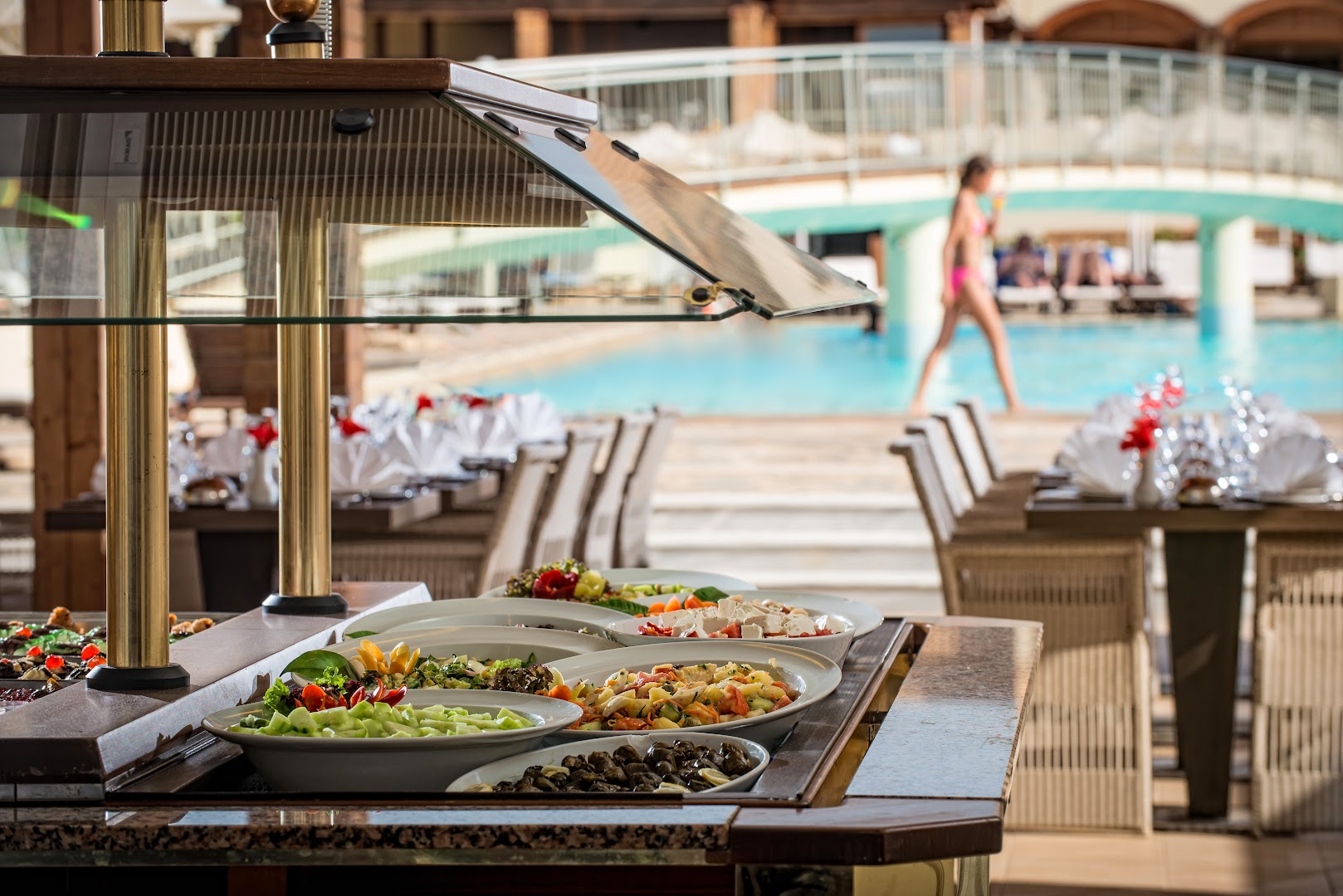 Bella Beach Hotel bufetes galds ar svaigiem salātiem un uzkodām, fonā redzams āra baseins un tilts Krētā