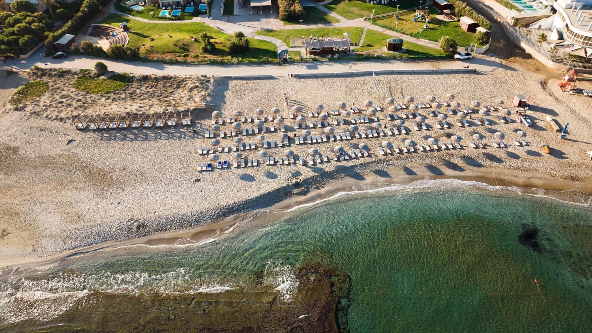 Bella Beach Hotel aerofoto skats uz plašo smilšu pludmali ar saulessargiem un dzidro Vidusjūras ūdeni