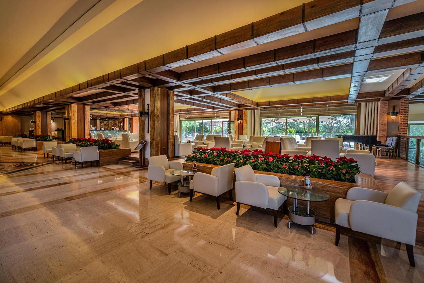 Gloria Golf Resort plašais lobby bārs ar ērtiem atpūtas krēsliem, ziedu dekorācijām un elegantiem koka griestiem