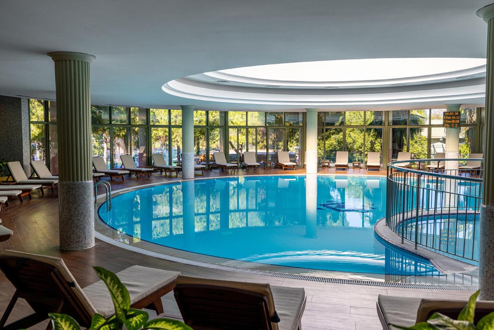 Gloria Golf Resort iekštelpu baseins ar panorāmas logiem, atpūtas krēsliem un dabisko apgaismojumu