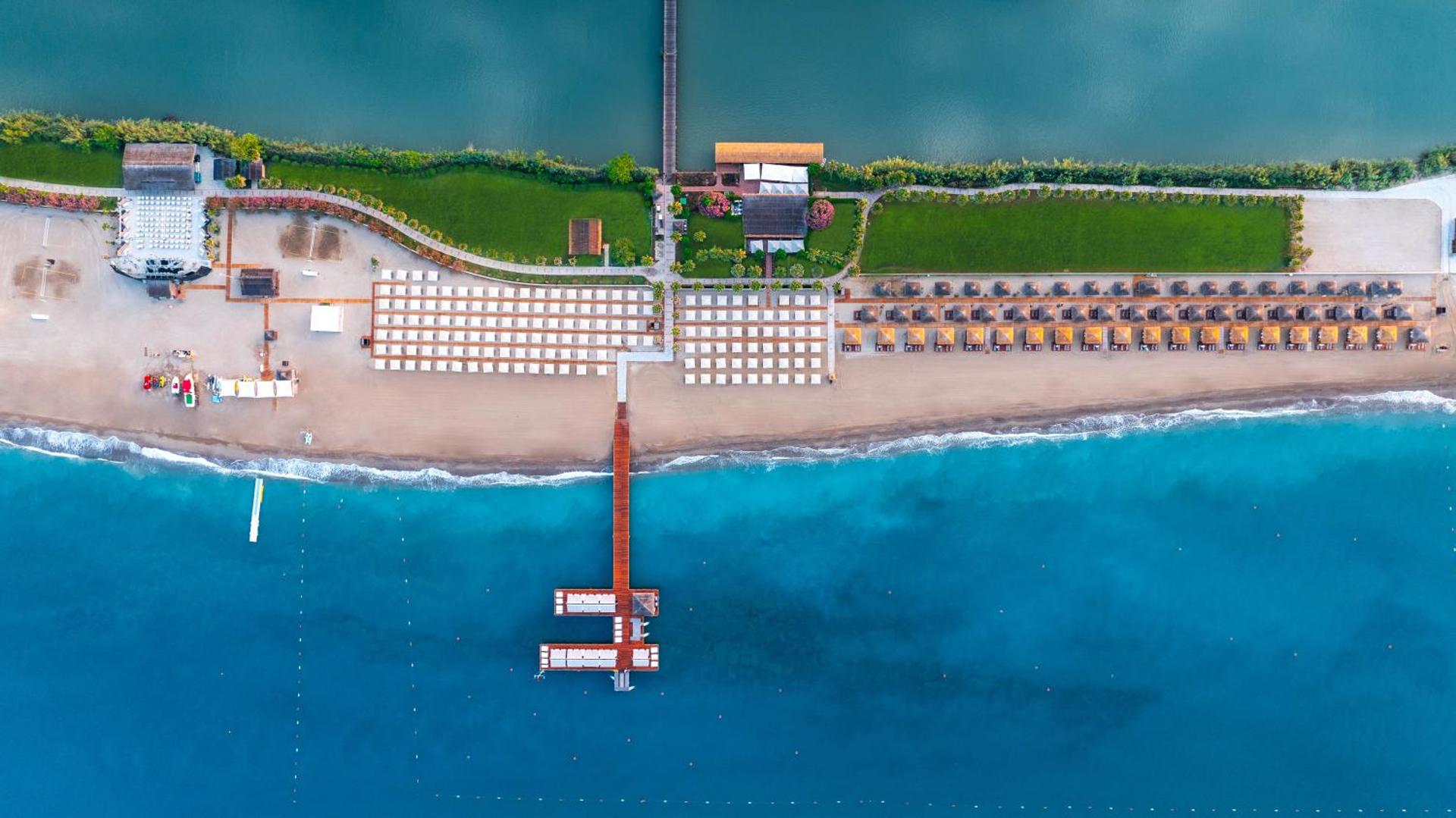 Gloria Golf Resort pludmales zonas aerofoto Belekā — plaša smilšu josla, sauļošanās krēsli, zaļie mauriņi un mols tirkīza krāsas ūdenī