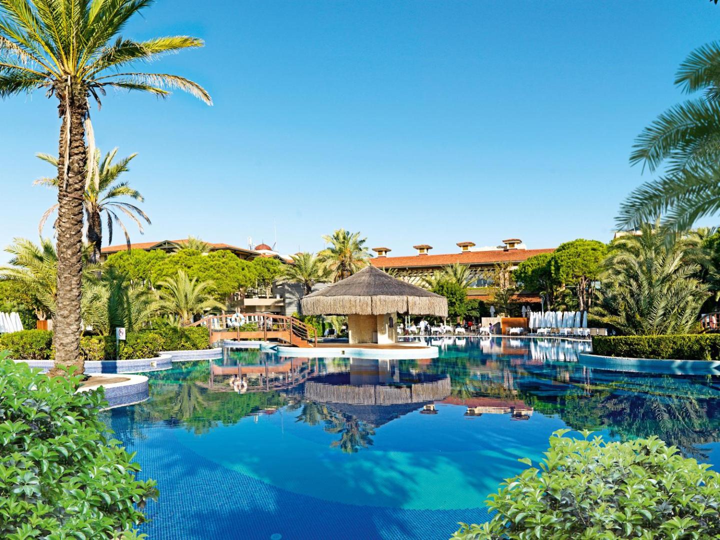 Gloria Golf Resort galvenais baseins ar bāru zem niedru jumta, palmām un viesnīcas ēku fonā saulainā dienā