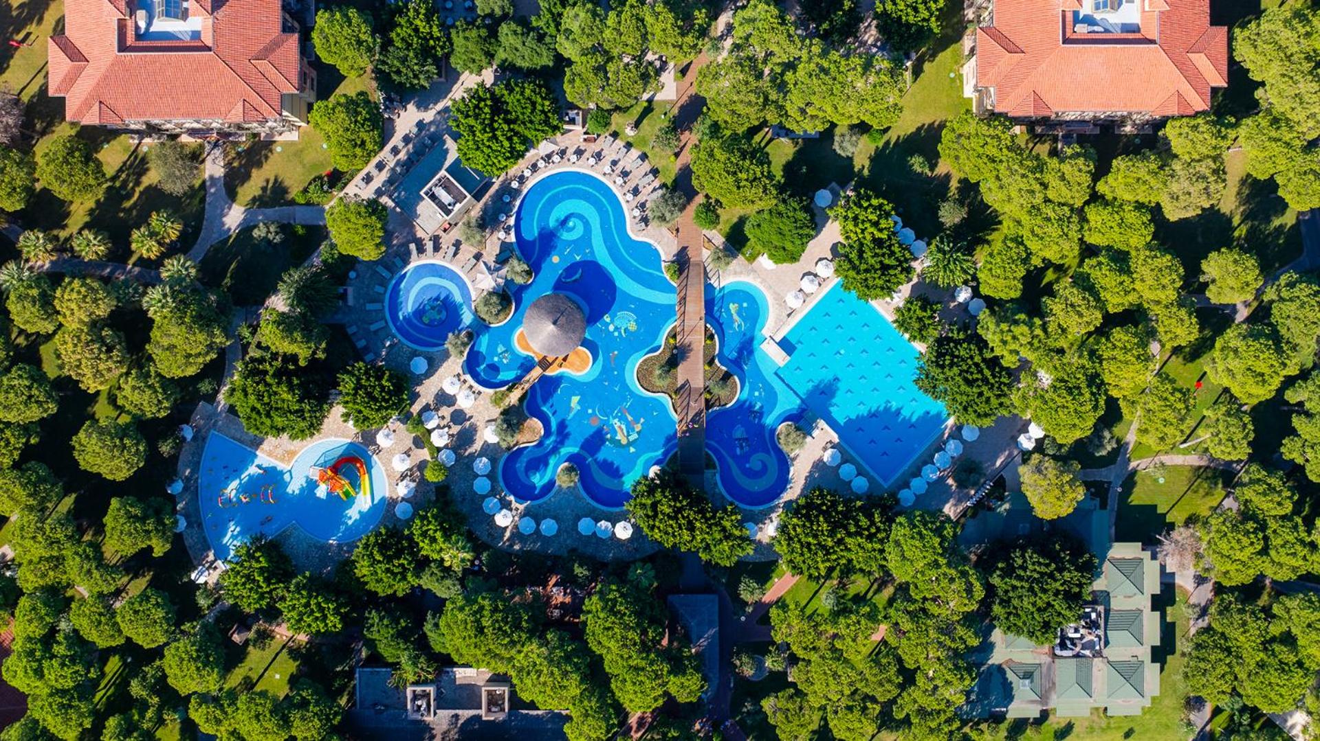 Gloria Golf Resort Belekā skats no putna lidojuma uz plašo baseinu kompleksu, ko ieskauj kupli dārzi un priežu mežs
