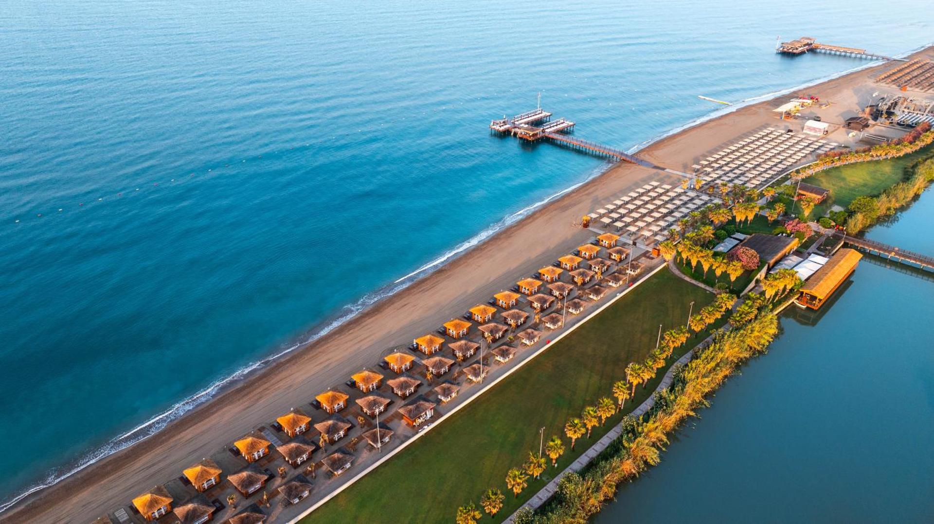 Gloria Golf Resort plašā smilšu pludmale Belekā ar privātām nojumēm, koka piestātni un skatu uz Vidusjūru saulrietā
