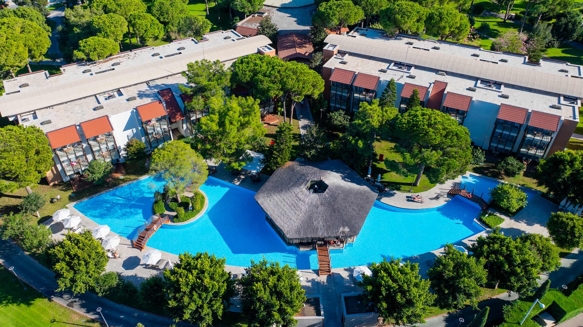 Gloria Golf Resort āra baseins ar bāru zem salmu jumta, ko ieskauj viesnīcas korpusi un zaļi koki Belekā