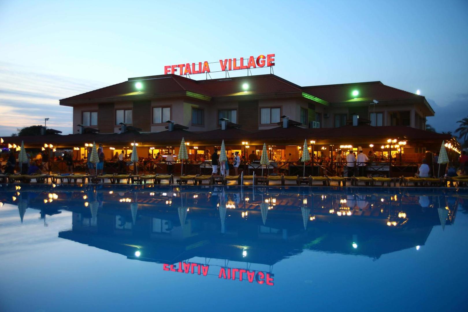 Eftalia Village galvenā ēka vakarā ar izgaismotu nosaukumu un restorāna terasi, kas atspīd mierīgā baseina ūdenī