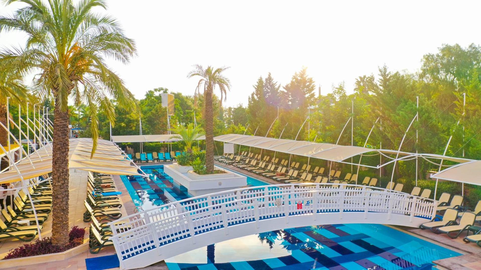Crystal Palace Luxury Resort & Spa āra baseins ar dekoratīvu tiltiņu, palmām un saulessargiem saulainā pēcpusdienā