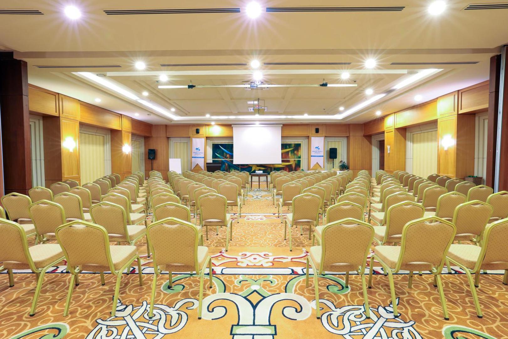 Plaša konferenču zāle Crystal Palace Luxury Resort & Spa viesnīcā Sidē — teātra stila sēdvietas, projektora ekrāns un grezns paklājs pasākumiem