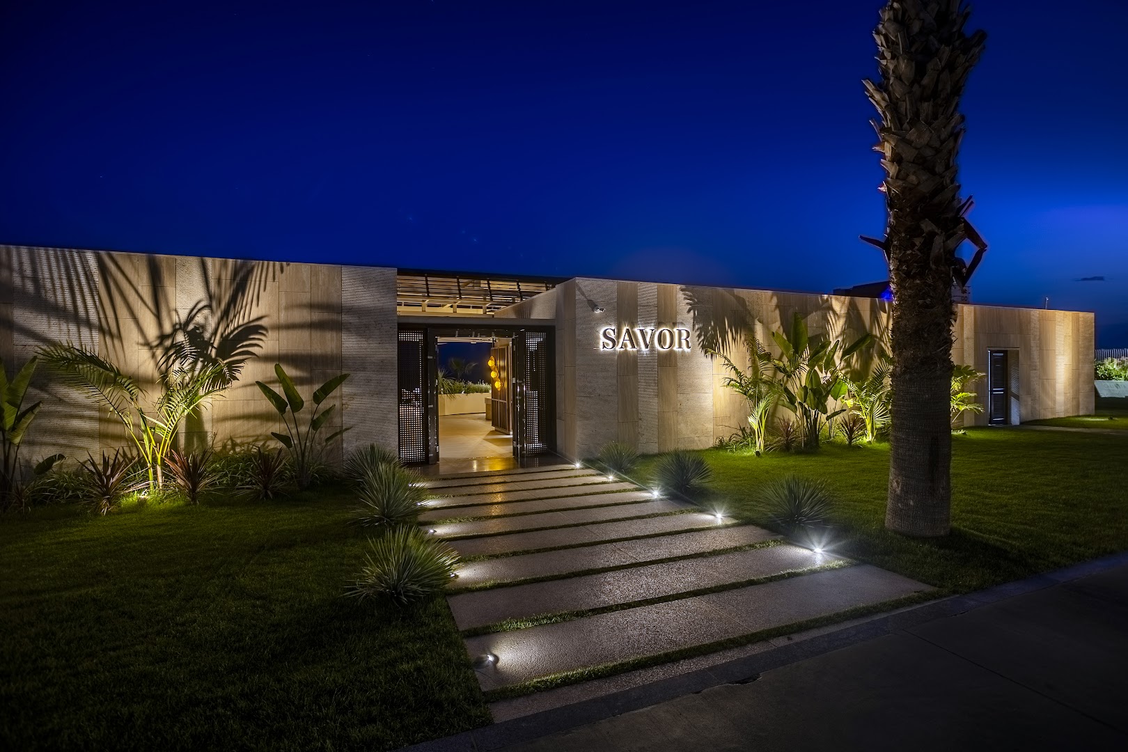 Calista Luxury Resort restorāna Savor ieeja vakarā ar izgaismotu taciņu, zaļu zālienu un palmām Belekā