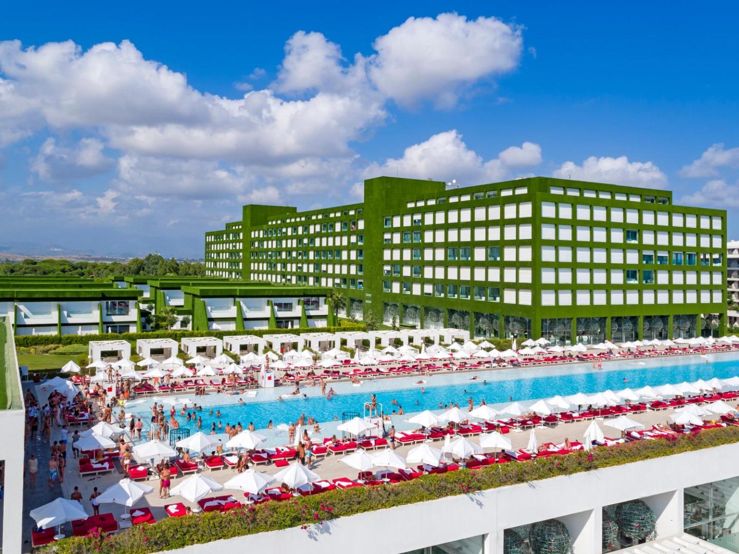 Adam & Eve Hotel galvenais baseins dienas laikā ar daudziem atpūtniekiem, baltiem saulessargiem un skatu uz viesnīcas zaļo galveno ēku