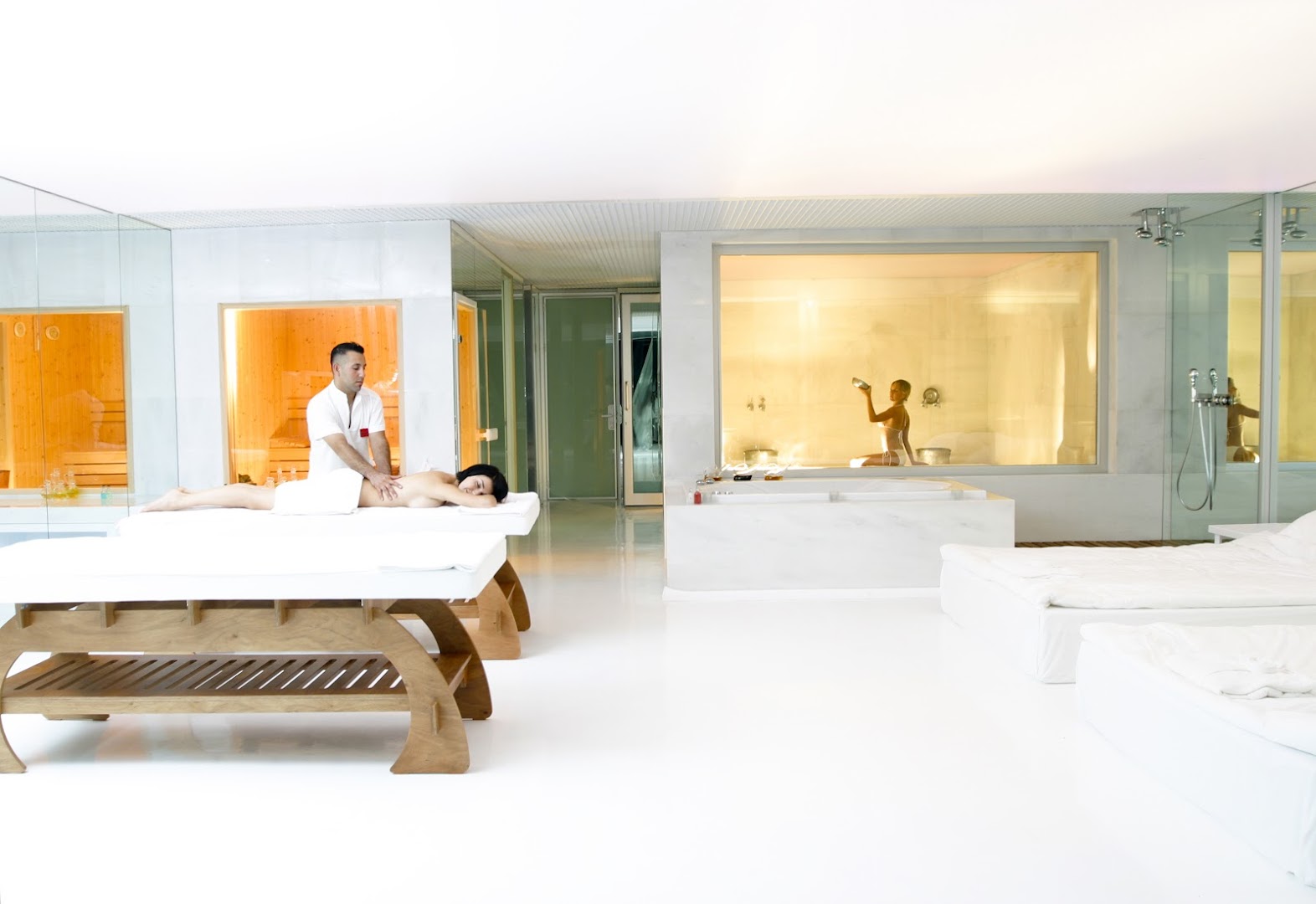 Adam & Eve Hotel spa centra masāžas telpa ar baltu dizainu, kurā redzama kliente un masieris darba procesā