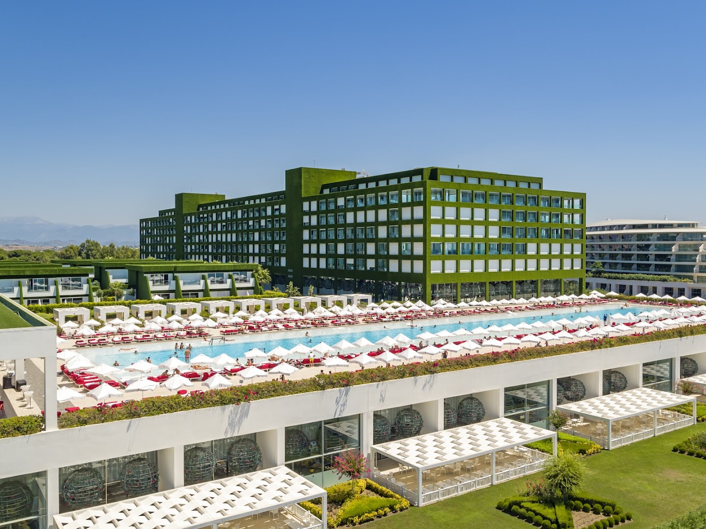 Adam & Eve Hotel modernā arhitektūra ar raksturīgo zaļo fasādi un garo āra baseinu, ko ieskauj balti saulessargi un atpūtas krēsli