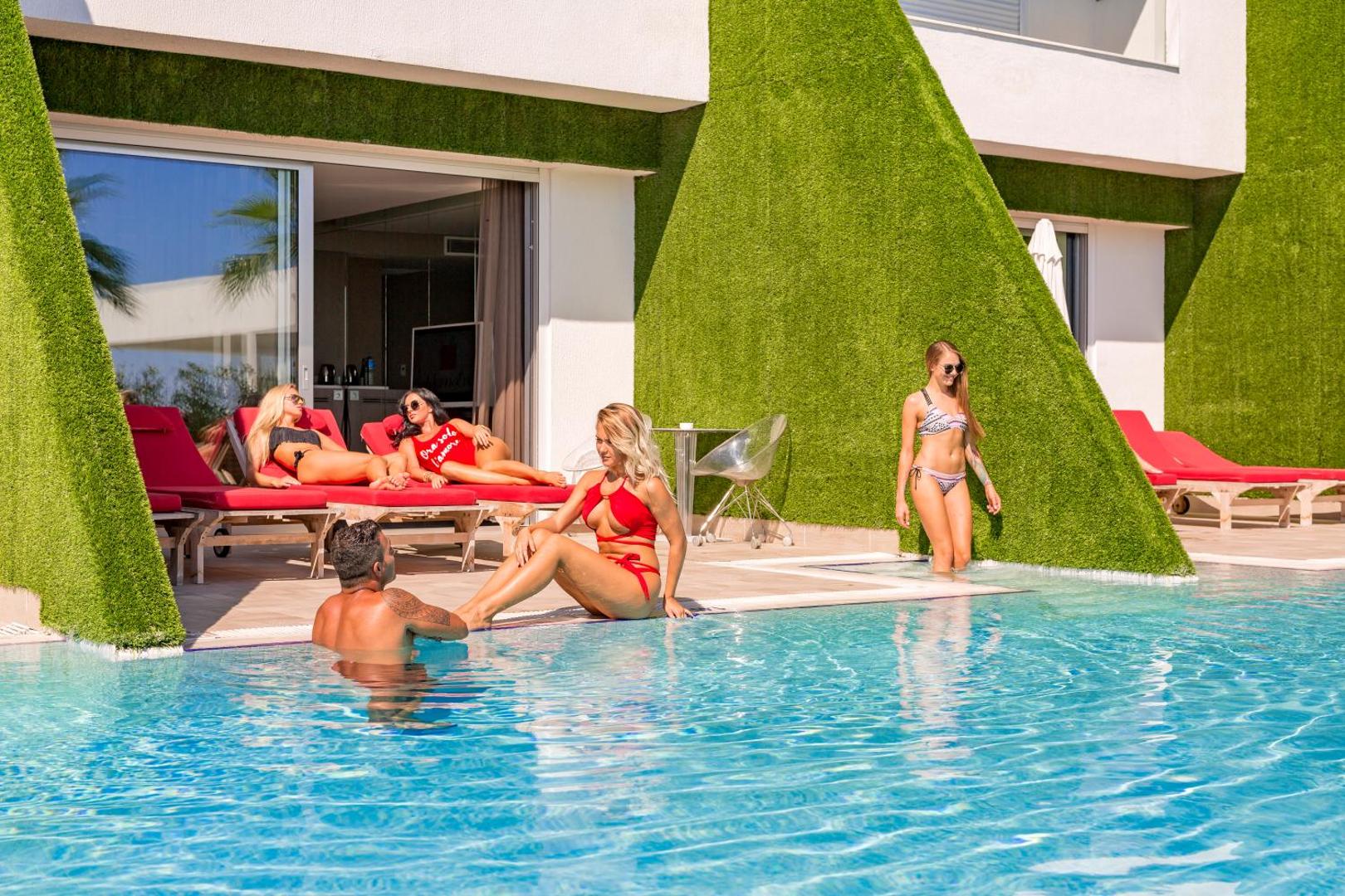 Adam & Eve Hotel dizaina numuri ar tiešu piekļuvi baseinam un terasi, kur izvietoti sarkani sauļošanās krēsli uz mākslīgā zālāja fona