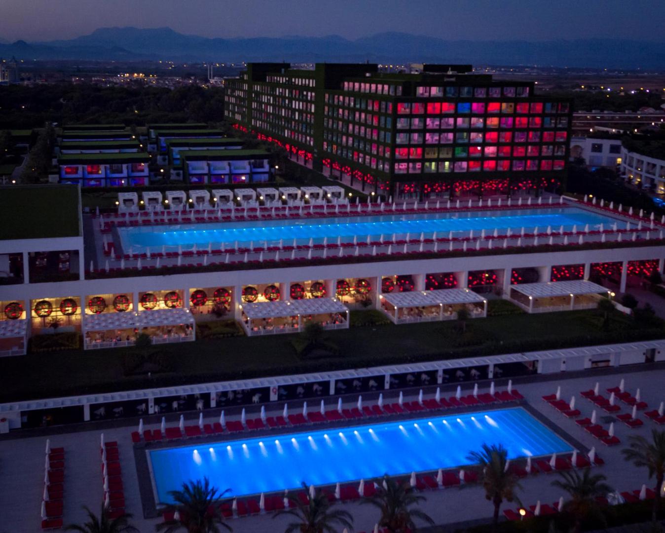 Adam & Eve Hotel nakts skats ar izgaismotiem baseiniem un krāsainām gaismām viesnīcas numuru logos uz kalnu fona Belekā