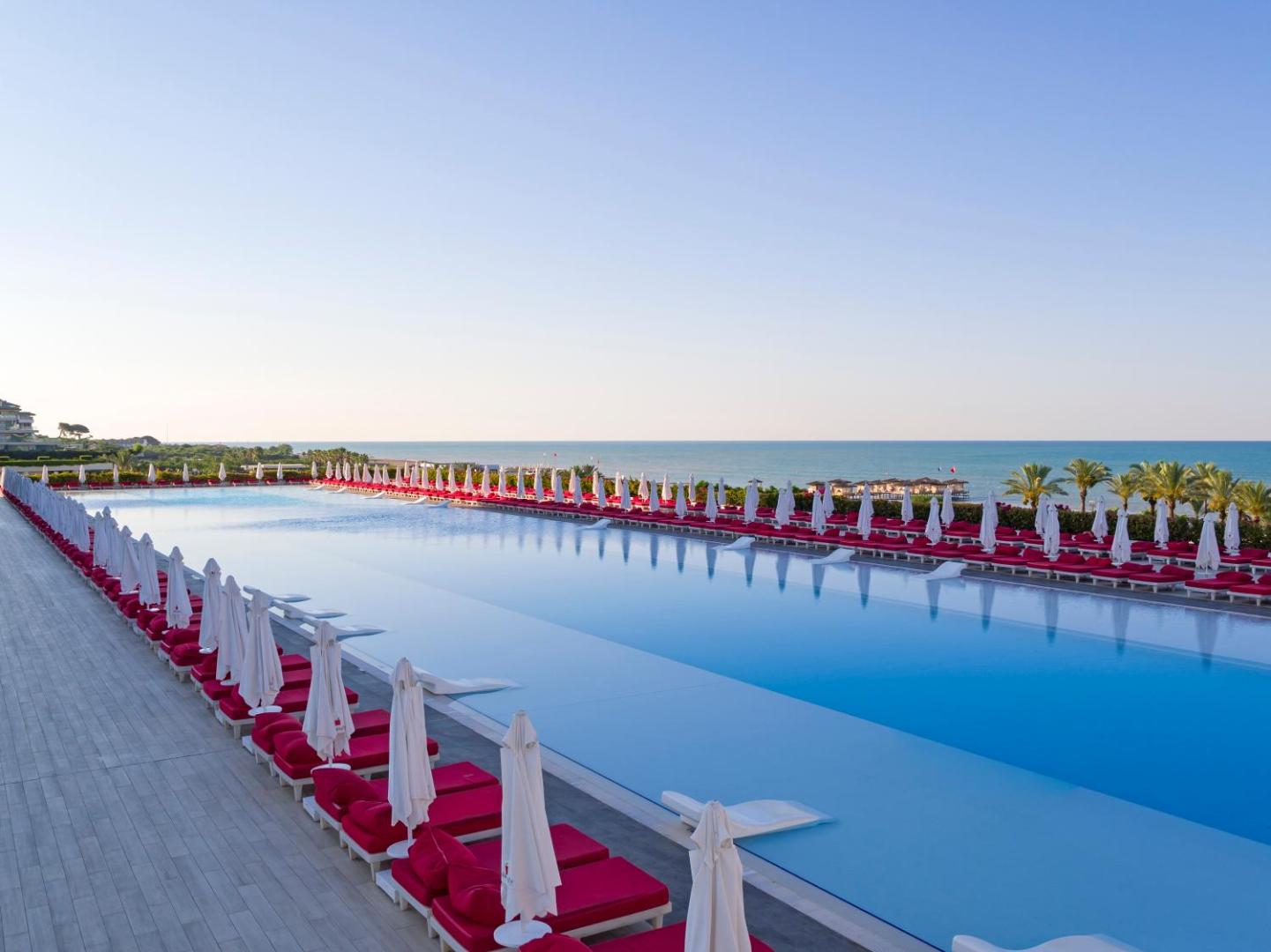 Adam & Eve Hotel bezgalības tipa baseins rīta gaismā ar rindu sarkanu sauļošanās krēslu un skatu uz tālo jūras horizontu