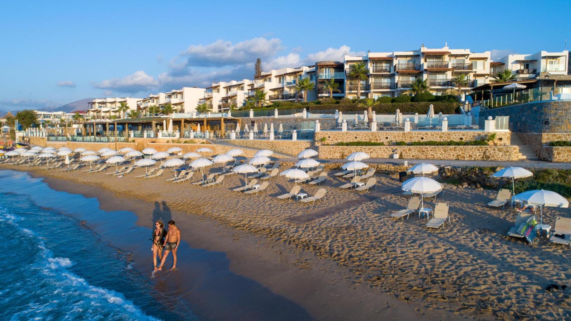 Alexander Beach Hotel & Village panorāmas skats uz smilšu pludmali ar saulessargiem un viesnīcas ēkām saulrietā Malijā, Krētā
