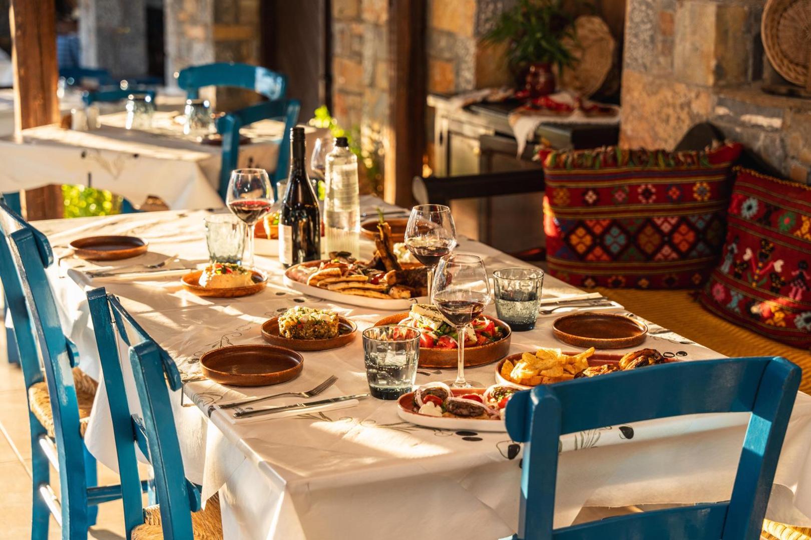 Galds ar tradicionāliem grieķu ēdieniem un vīnu Alexander Beach Hotel & Village tavernā, fonā akmens sienas un zili krēsli