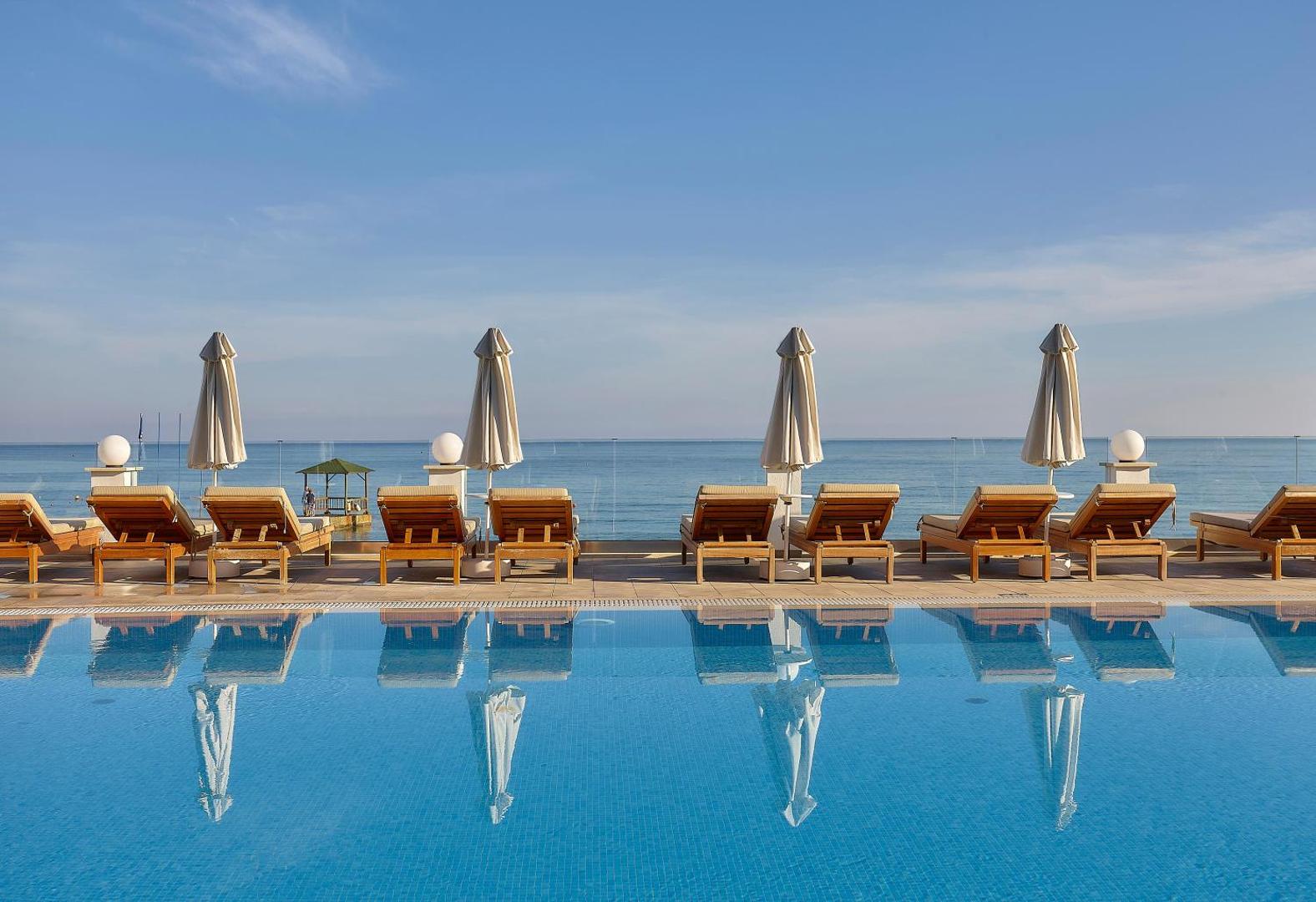 Alexander Beach Hotel & Village bezgalības tipa baseins ar koka sauļošanās krēsliem un saulessargiem tieši pie zilās jūras malas