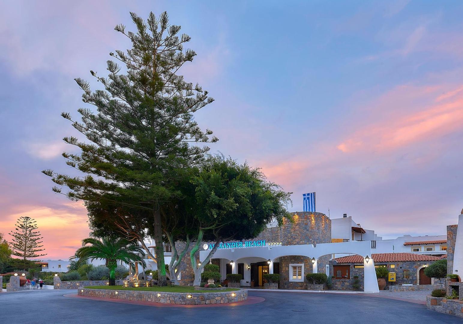 Alexander Beach Hotel & Village galvenā ieeja vakarā ar izgaismotu logo, lielu priedi un tradicionālu akmens arhitektūru.