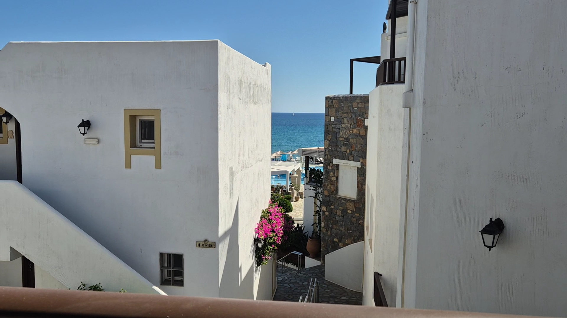 Skats starp baltām Alexander Beach Hotel & Village ēkām uz zilo jūru, ziedošiem bugenvilleju krūmiem un akmens sienām.