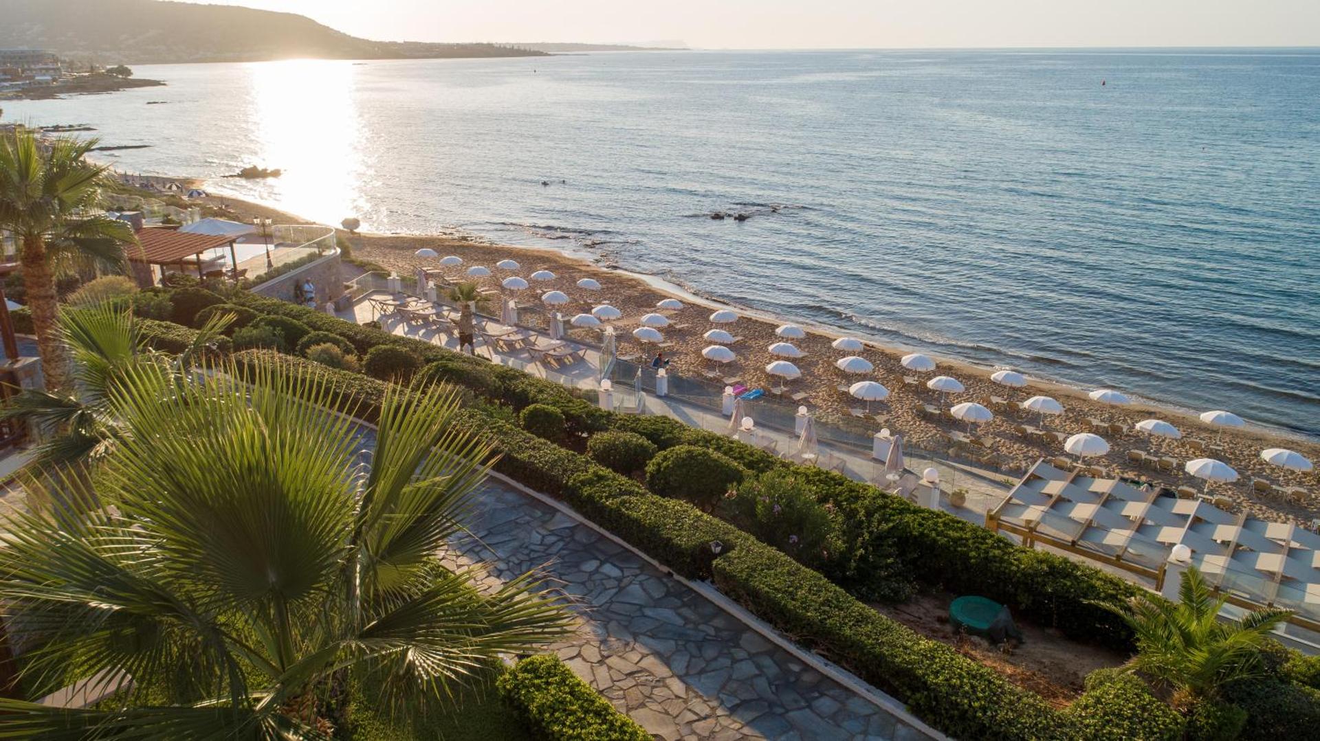 Alexander Beach Hotel & Village plašā smilšu pludmale ar saulessargiem un koptu dārzu piekrastē saules apspīdētā pēcpusdienā