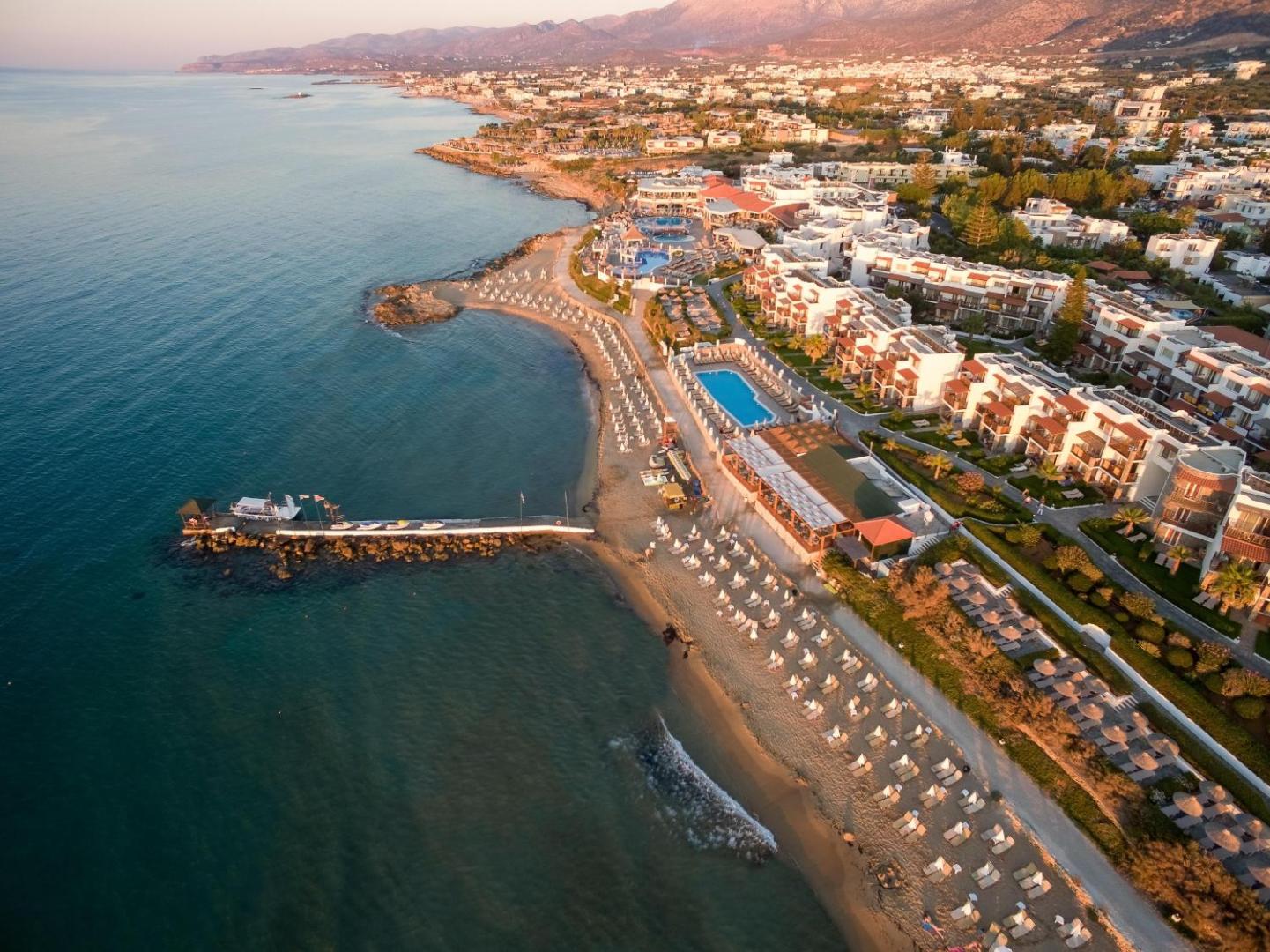 Alexander Beach Hotel & Village aerofotoattēls saulrietā — plaša smilšu pludmale, piestātne un viesnīcas korpusi pie jūras Malijā, Krētā.
